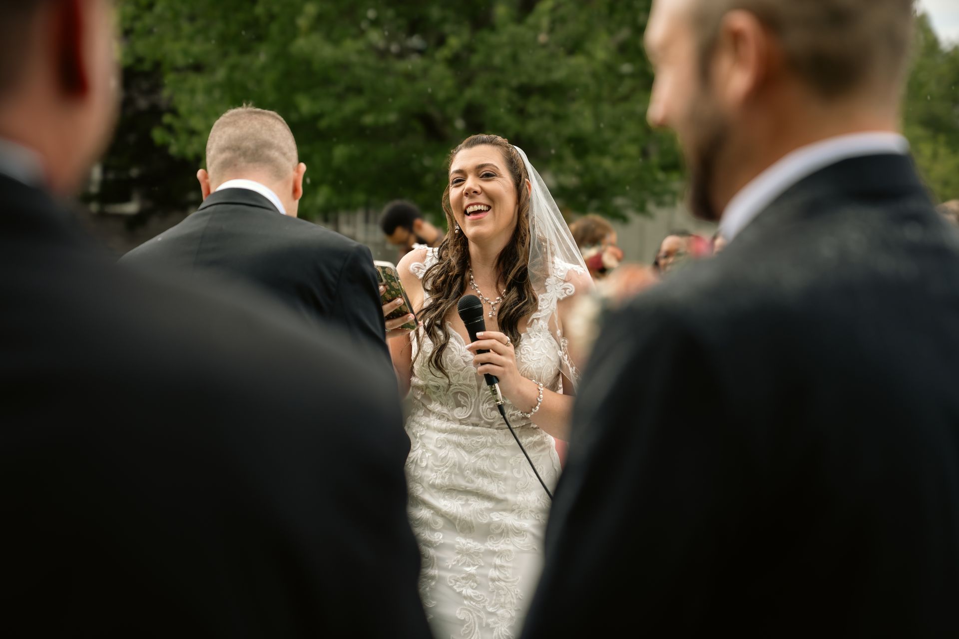 A bride in a white lace dress and veil holds a microphone and laughs during an outdoor wedding speech.