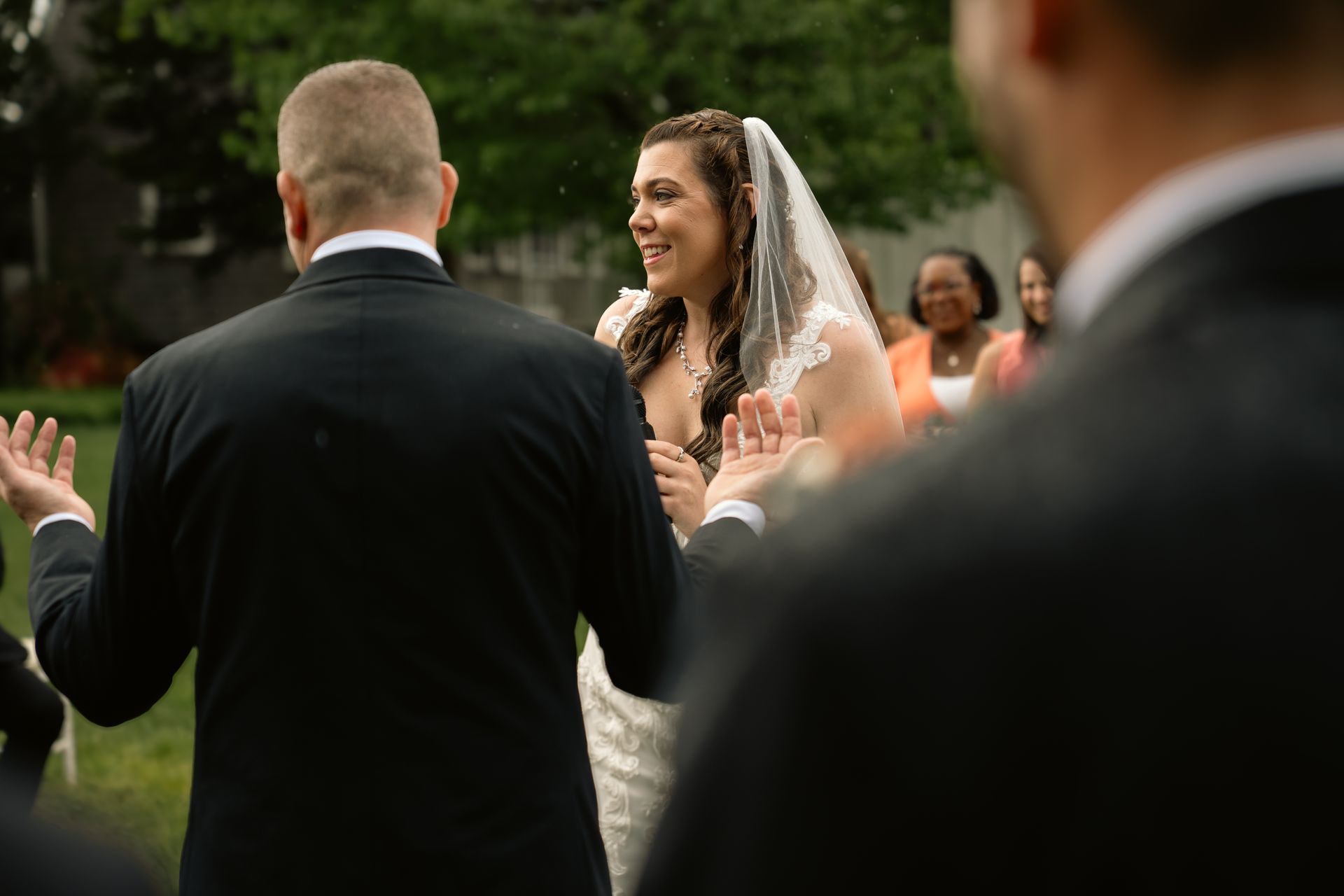 A groom facing his bride during an outdoor wedding ceremony, with guests blurred in the background.