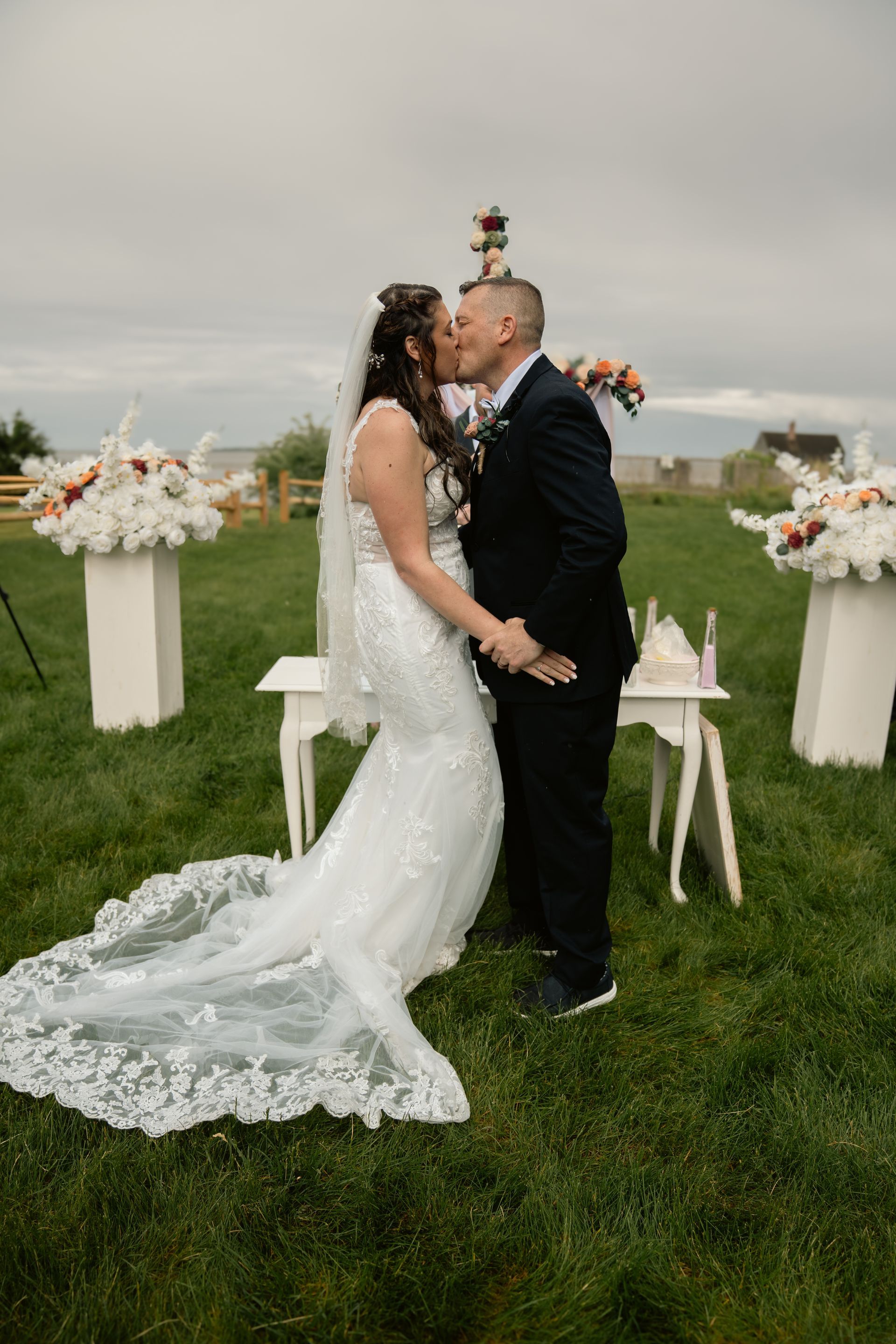 A couple kisses at an outdoor wedding ceremony, standing on grass between two floral arrangements on pedestals.