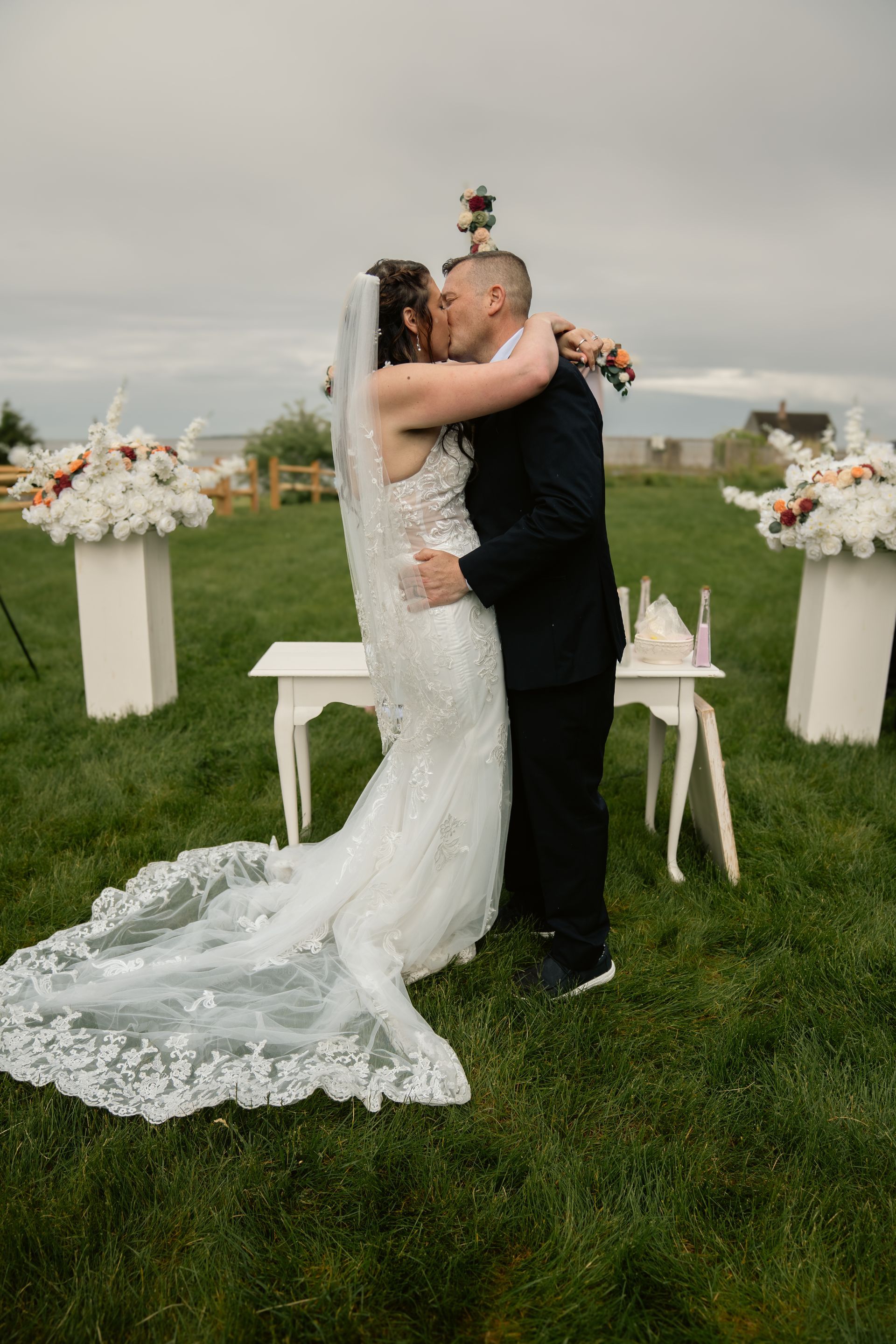 A couple kisses at an outdoor wedding ceremony in a grassy field, with floral arrangements on pedestals behind them.