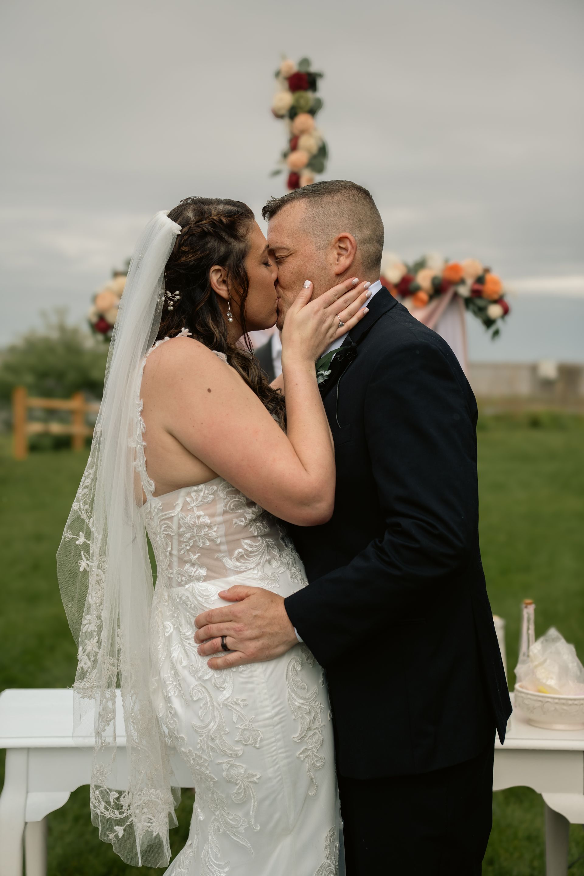 A bride and groom in wedding attire kiss during an outdoor ceremony in a grassy field.
