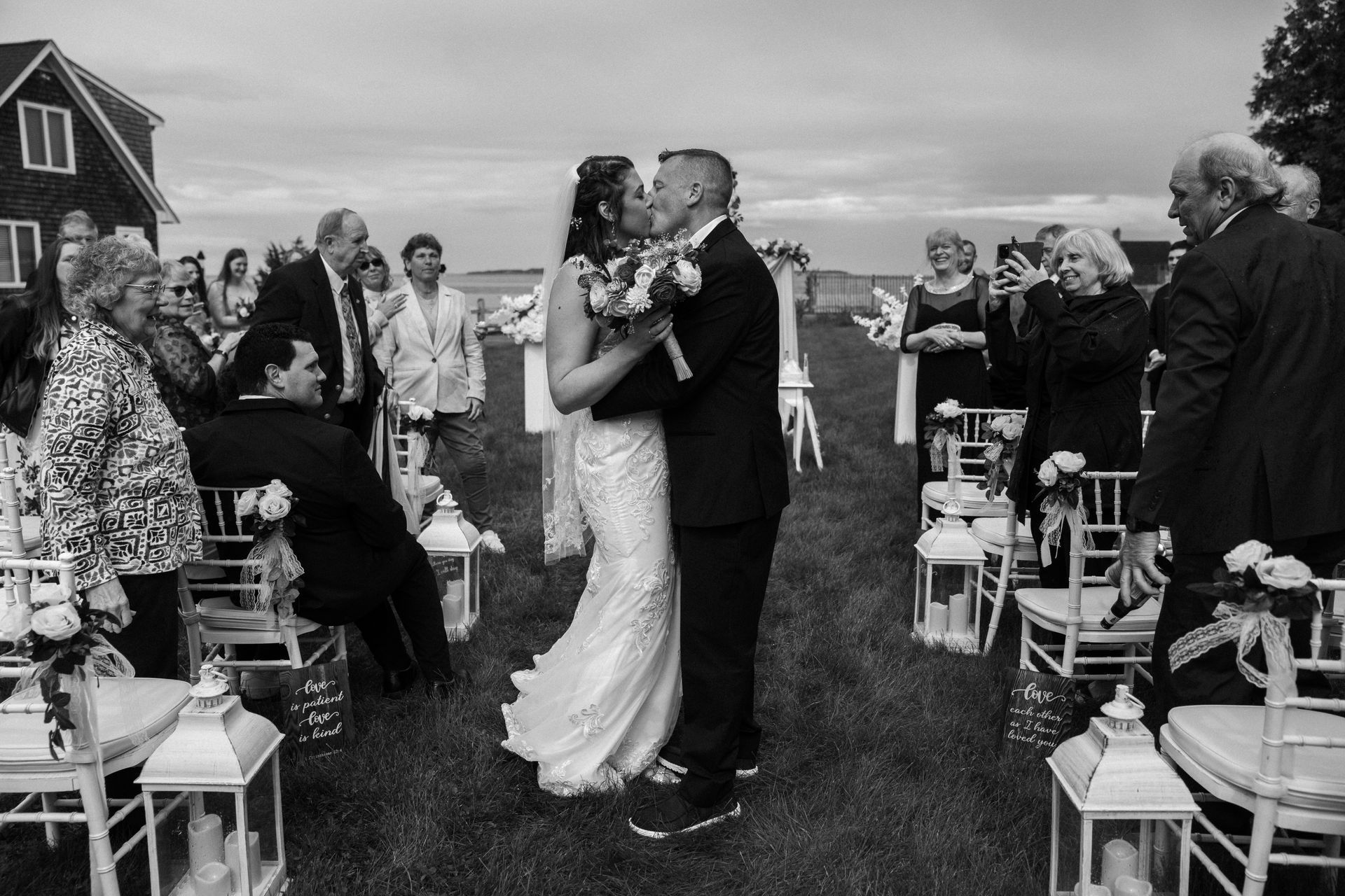 A couple kisses at their outdoor wedding ceremony, surrounded by seated guests and lanterns in a grassy field.