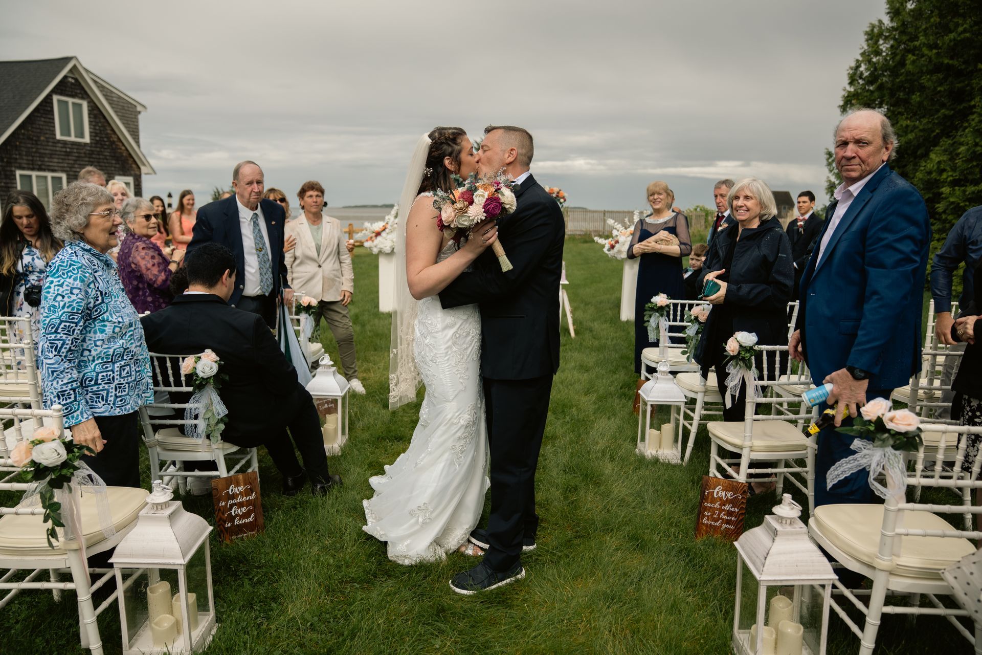 A bride and groom kiss at an outdoor wedding ceremony while guests seated on chairs watch from the grass.