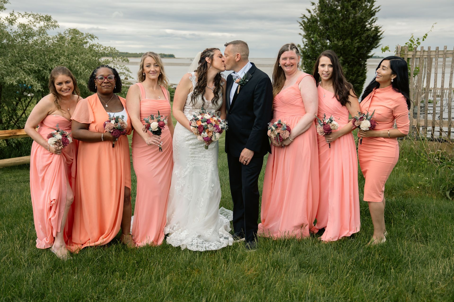 A wedding party stands outside, the bride and groom kissing, flanked by six bridesmaids in matching coral gowns.