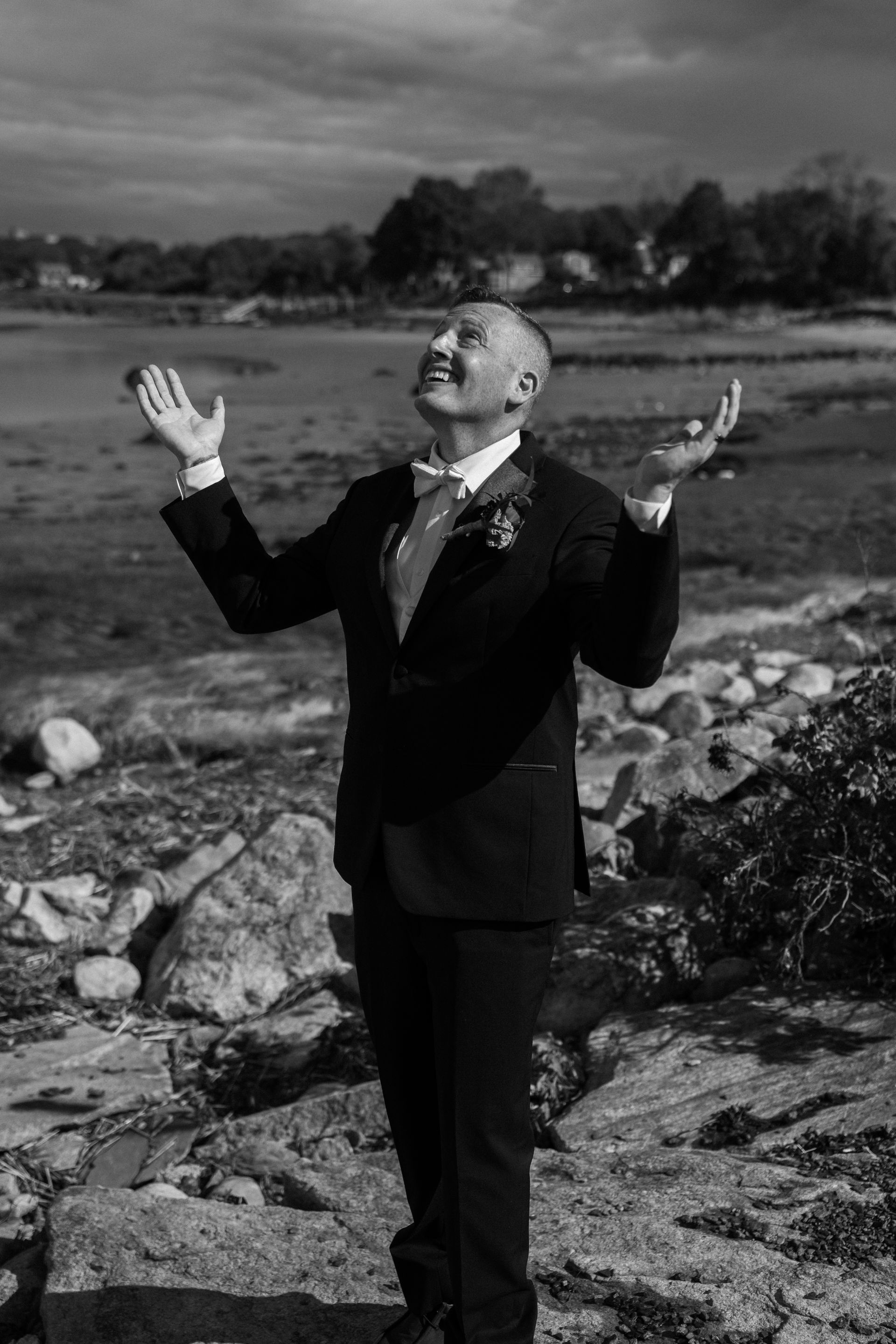 A person in a suit stands on a rocky beach with their arms outstretched and face turned upward toward the sky.