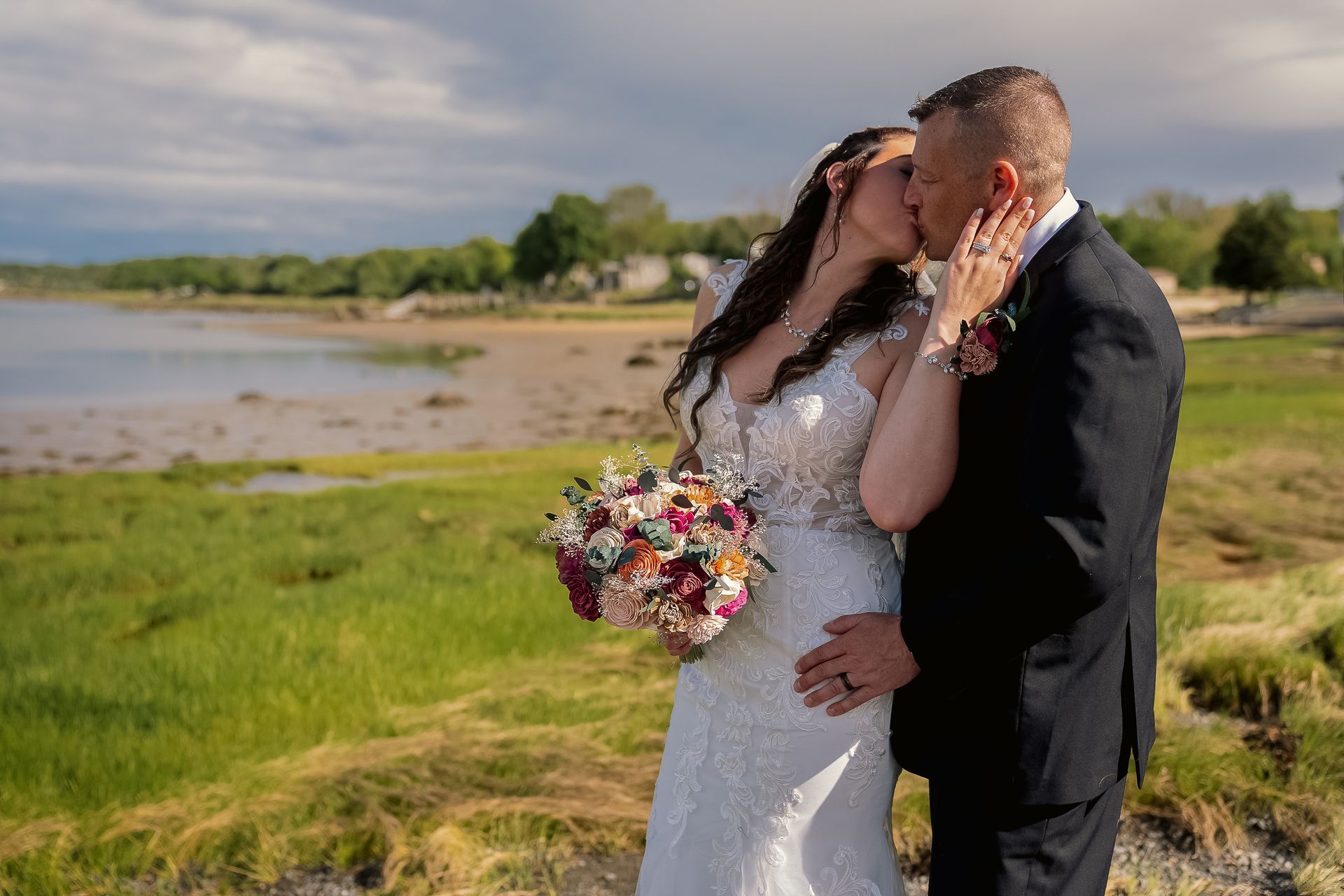 A bride in a lace gown and a groom in a suit kiss while standing on a grassy shore by the water.