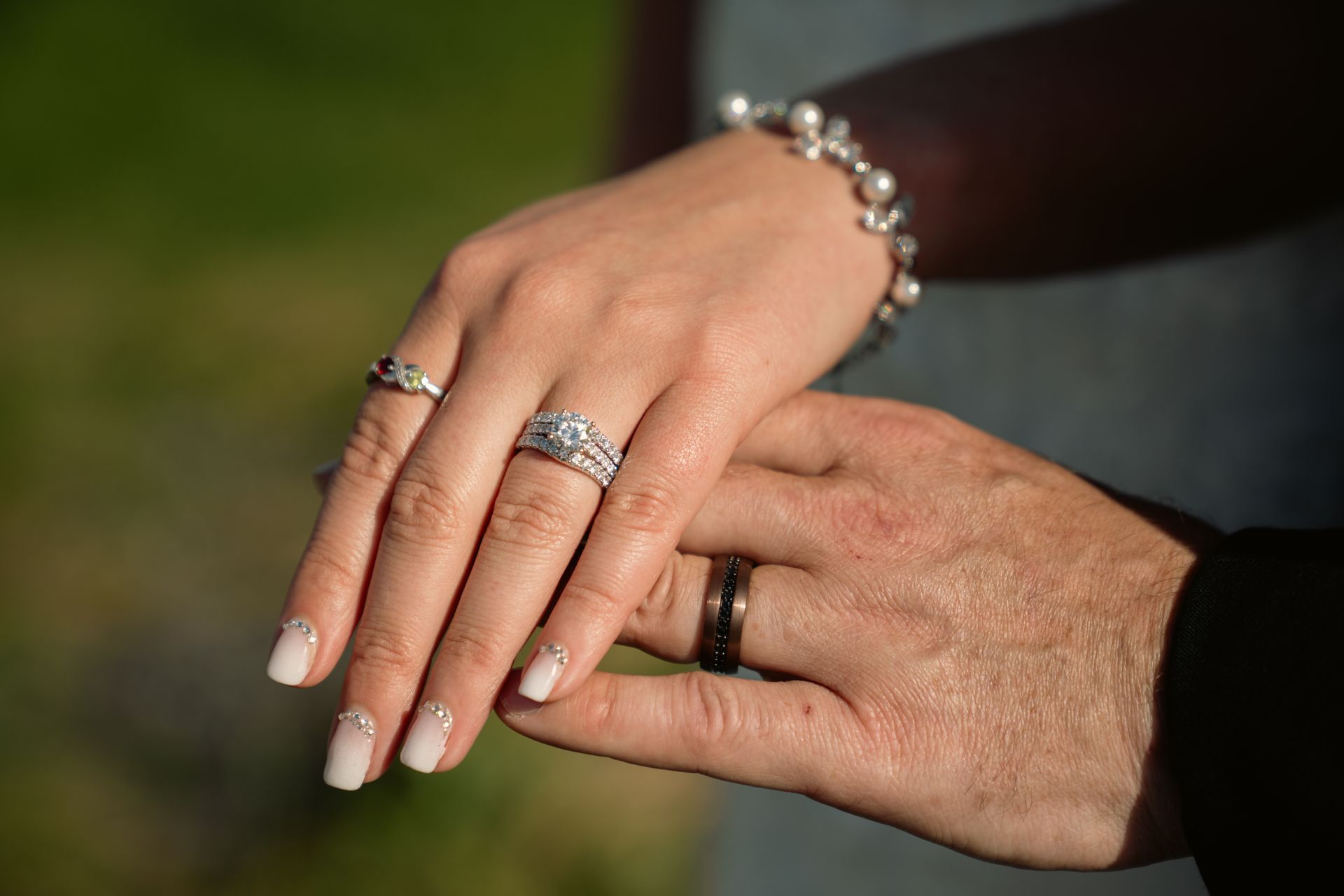 A close-up of two hands wearing wedding bands, one with a diamond ring and pearl bracelet, set against a blurred background.