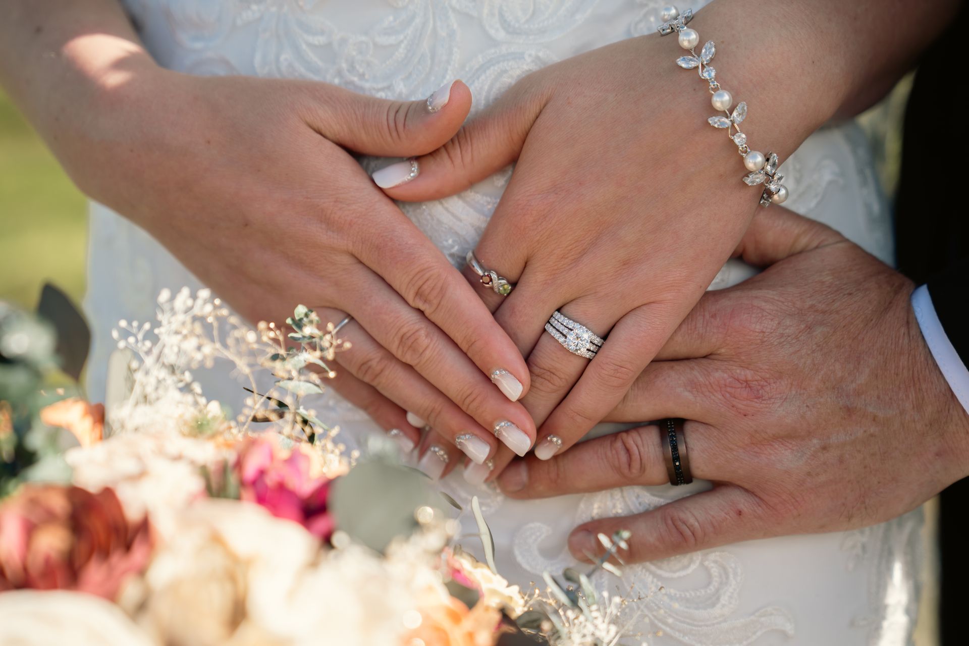 A couple’s hands joined over a wedding dress, showing wedding rings, a diamond engagement ring, and a floral bouquet.