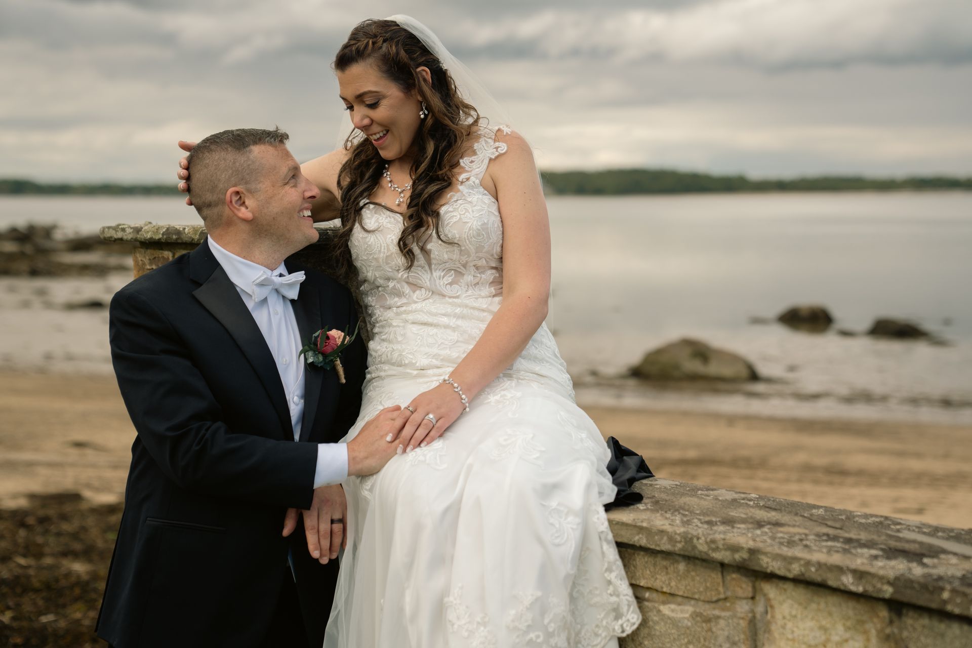 A bride in a white wedding dress sits on a stone wall by a lake, smiling at her groom who is kneeling beside her.
