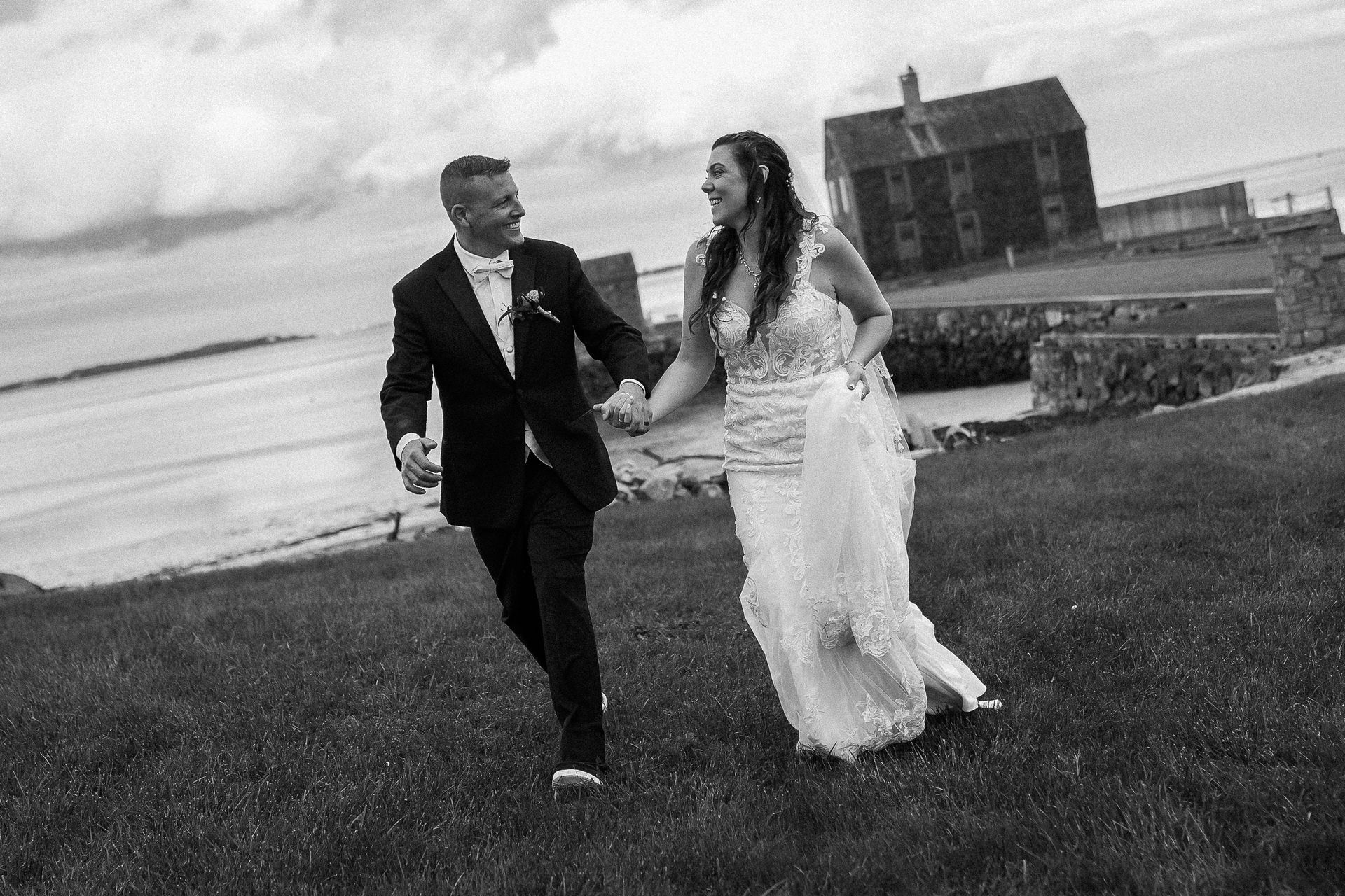 A couple in formal wedding attire holding hands and running across a grassy field near the ocean and a stone building.