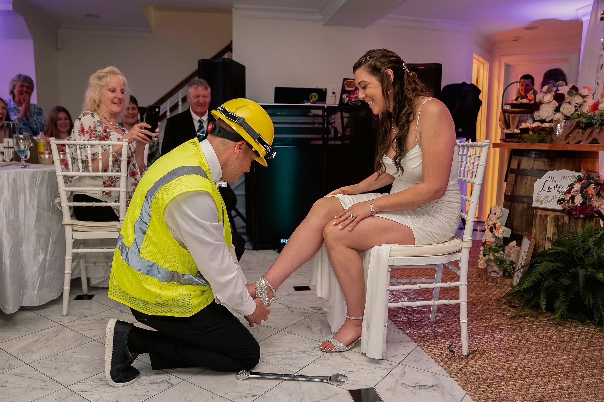 A person in a high-visibility vest and yellow hard hat kneels to put a shoe on a person sitting on a chair at an event.