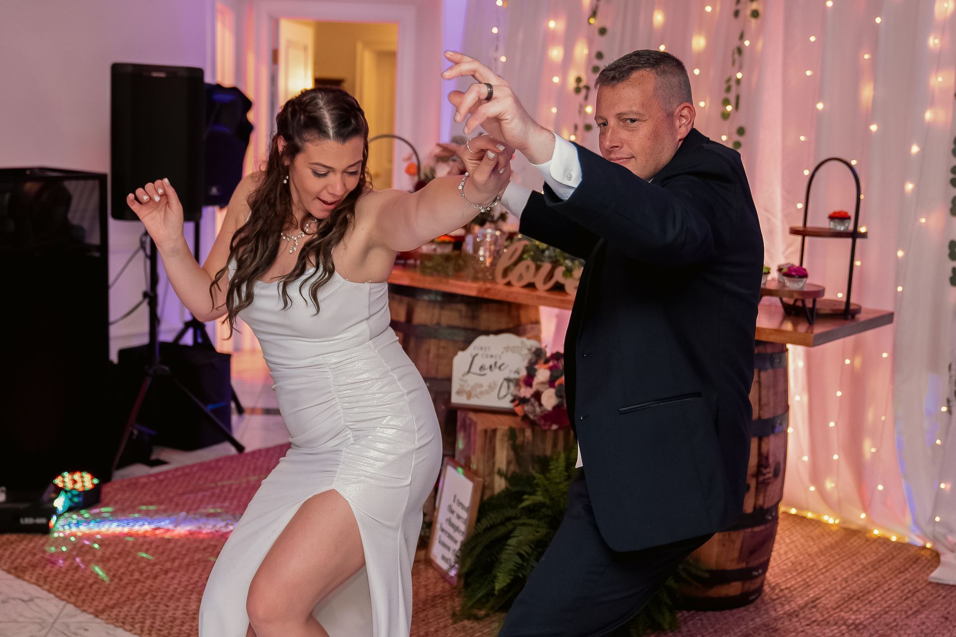 A bride and groom dancing together joyfully at an indoor wedding reception with warm lighting and a rustic barrel table.