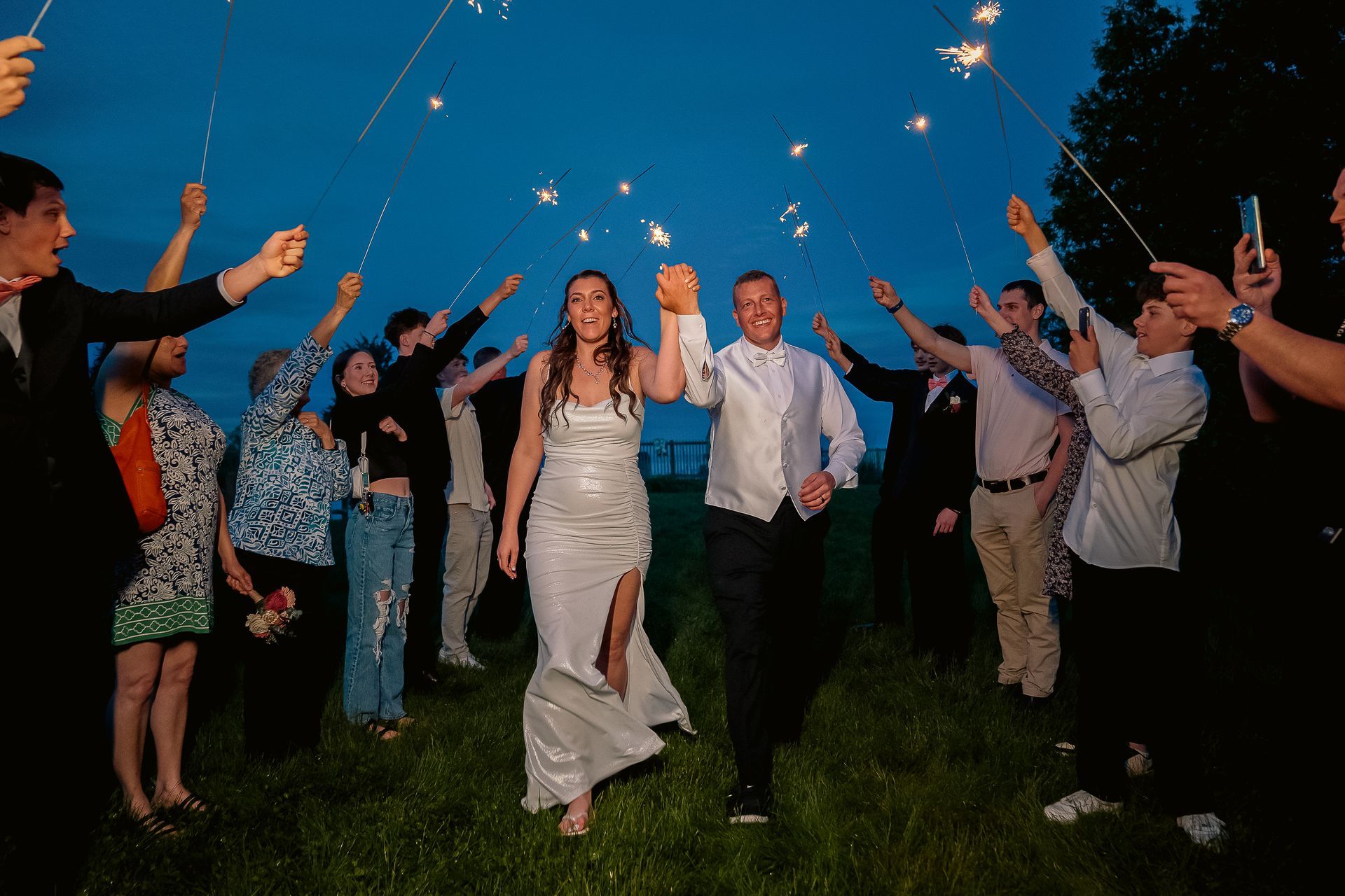 A bride and groom walk through a sparkler exit surrounded by guests at dusk in a grassy outdoor setting.