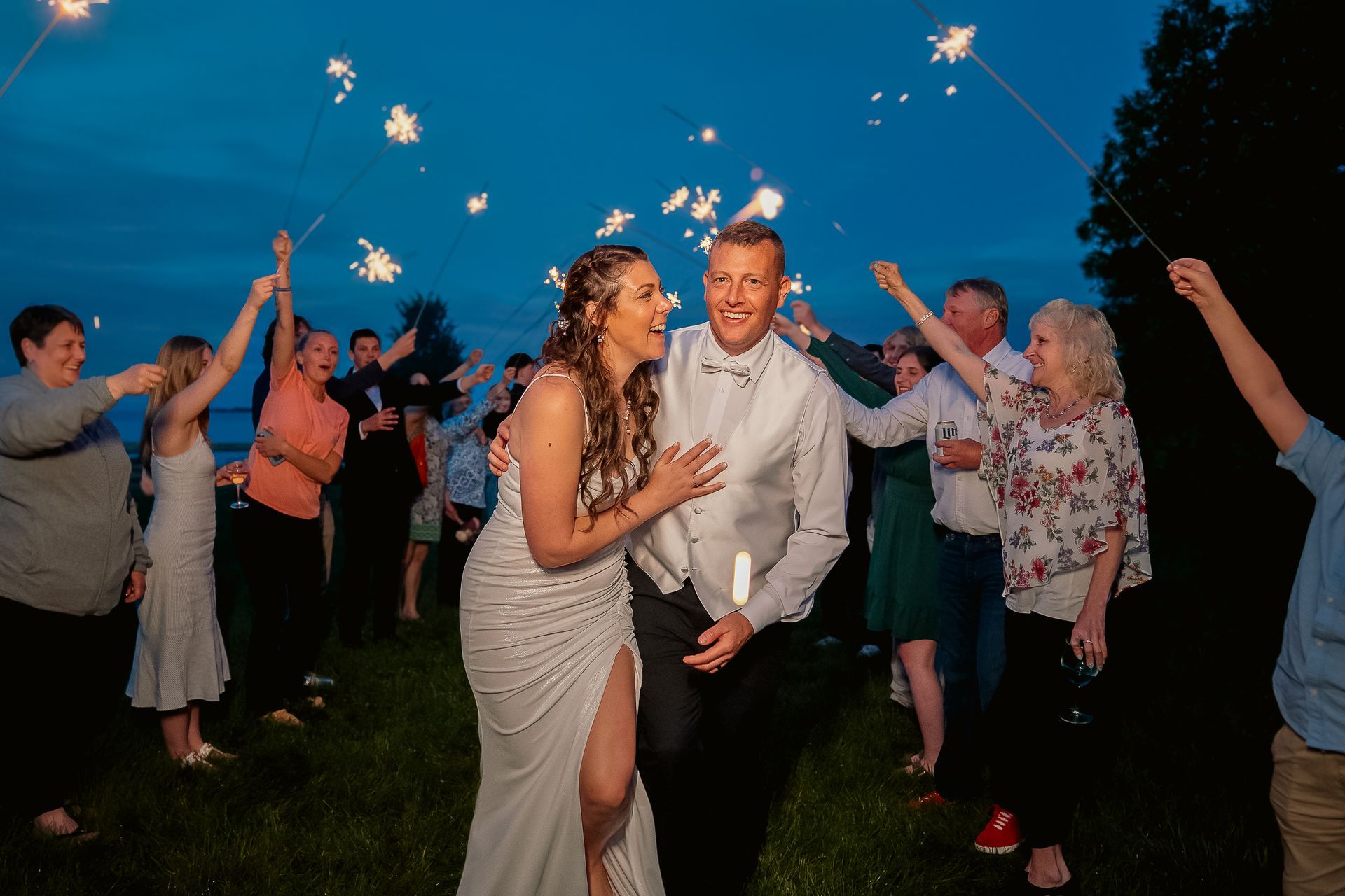 A bride and groom walk through a path of guests holding lit sparklers during an evening outdoor wedding celebration.