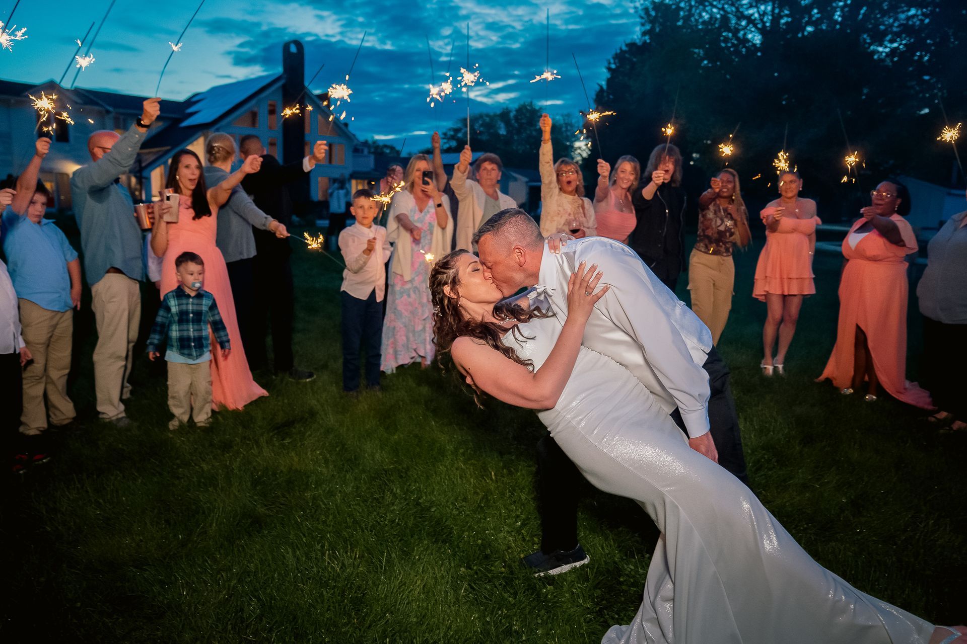 A bride and groom kiss while dipping backward as guests with sparklers surround them in an outdoor evening setting.