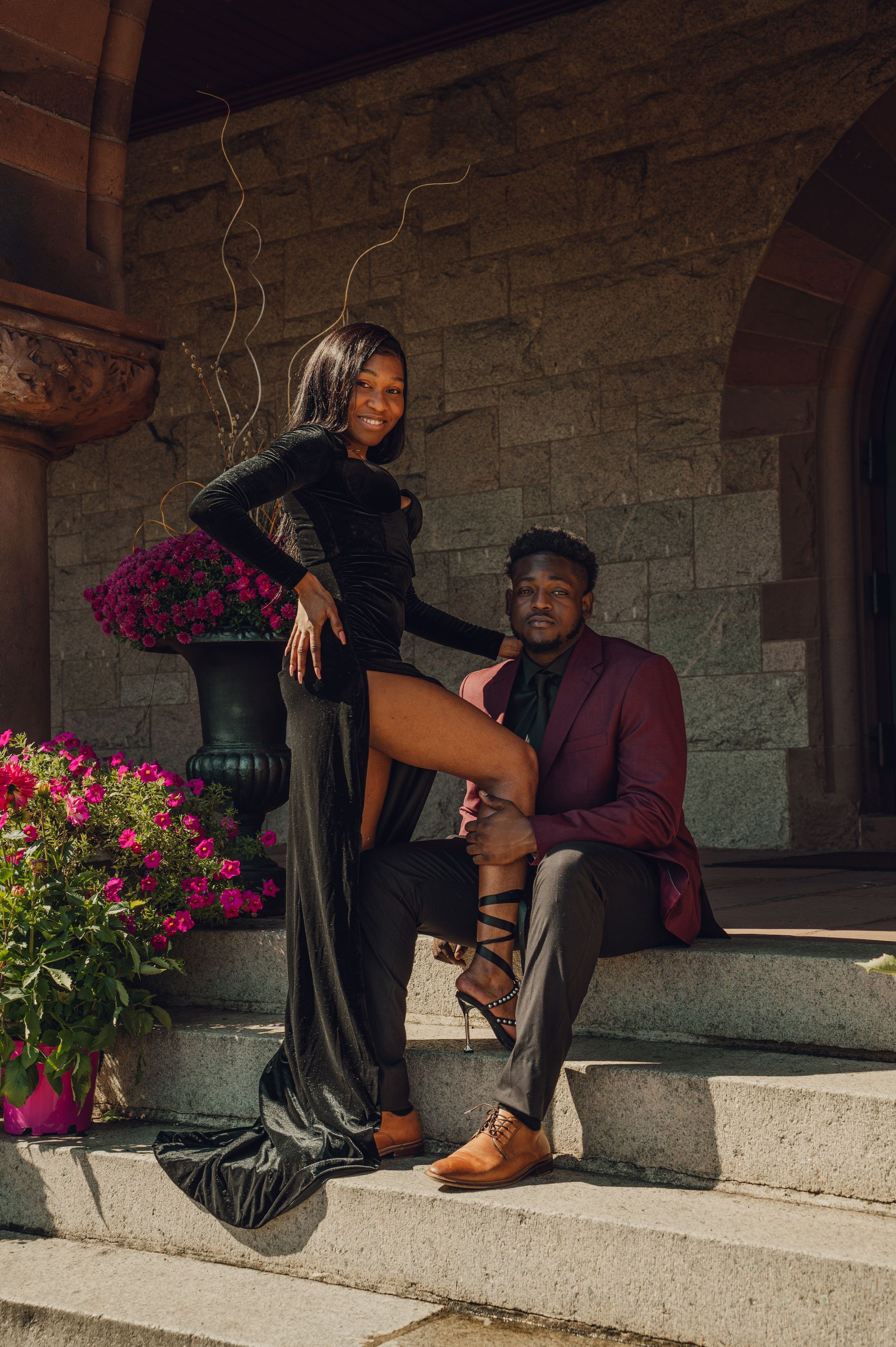 A couple in formal attire posing on stone steps in front of a stone building with decorative purple flowers.