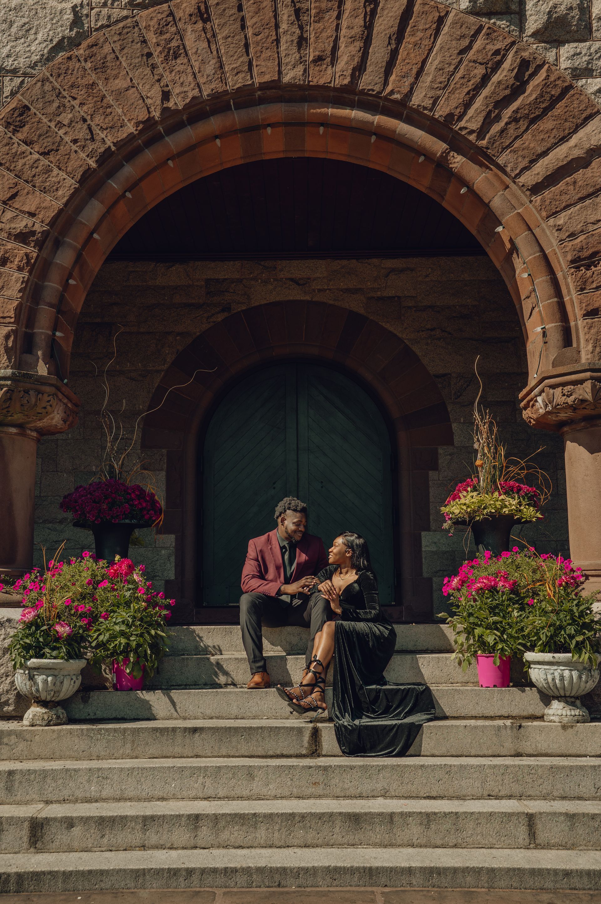 A couple sits on stone steps beneath a large stone archway, surrounded by potted flowers.
