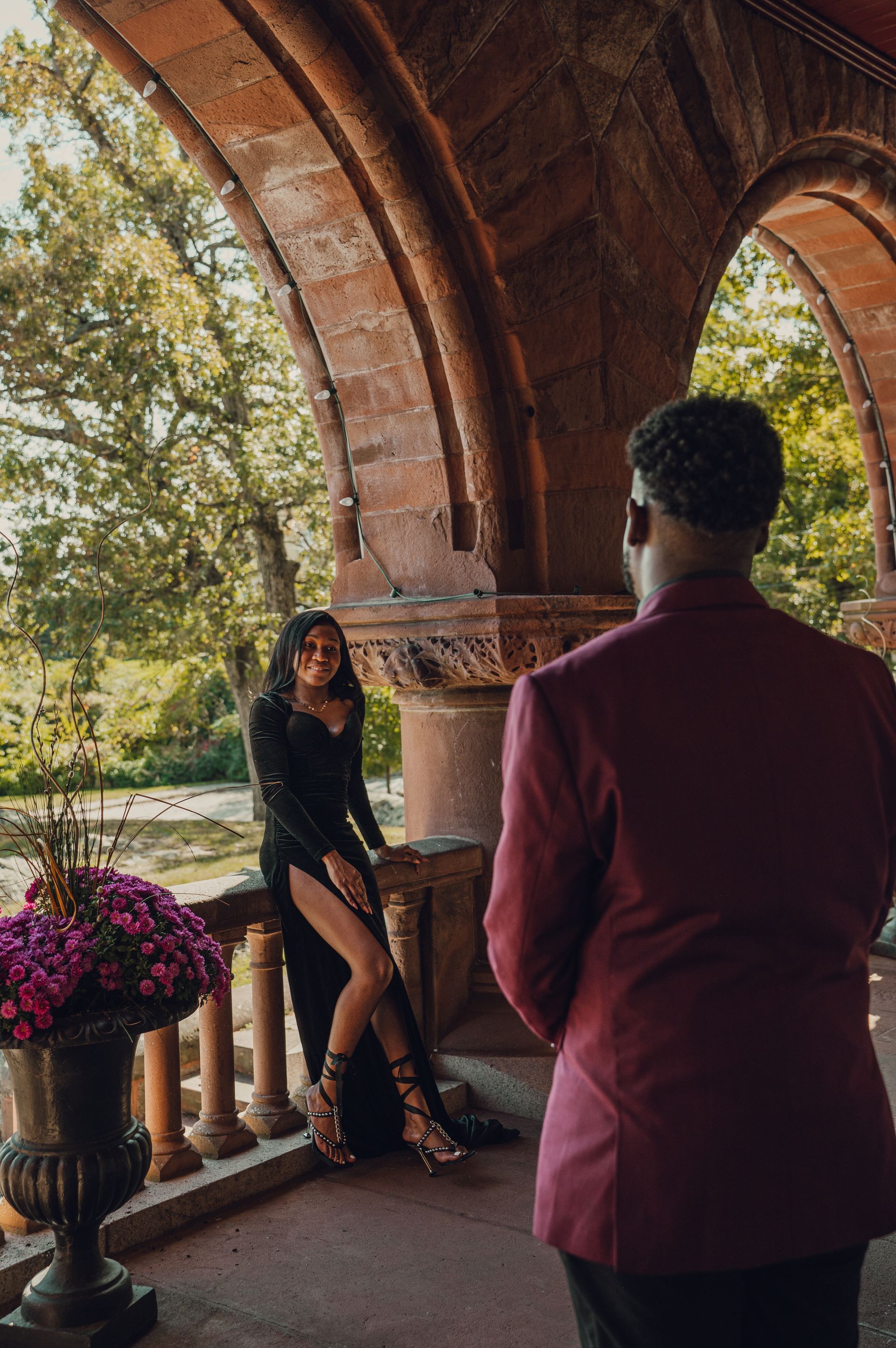 A person in a black gown poses on a stone balcony while another person in a maroon suit stands in the foreground.