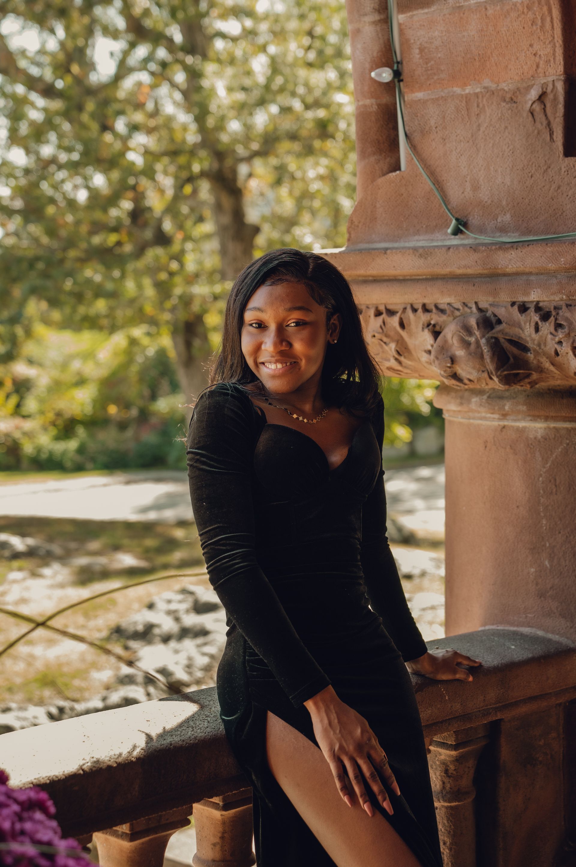 A smiling person in a long black dress leans against a stone railing outdoors with trees in the blurred background.