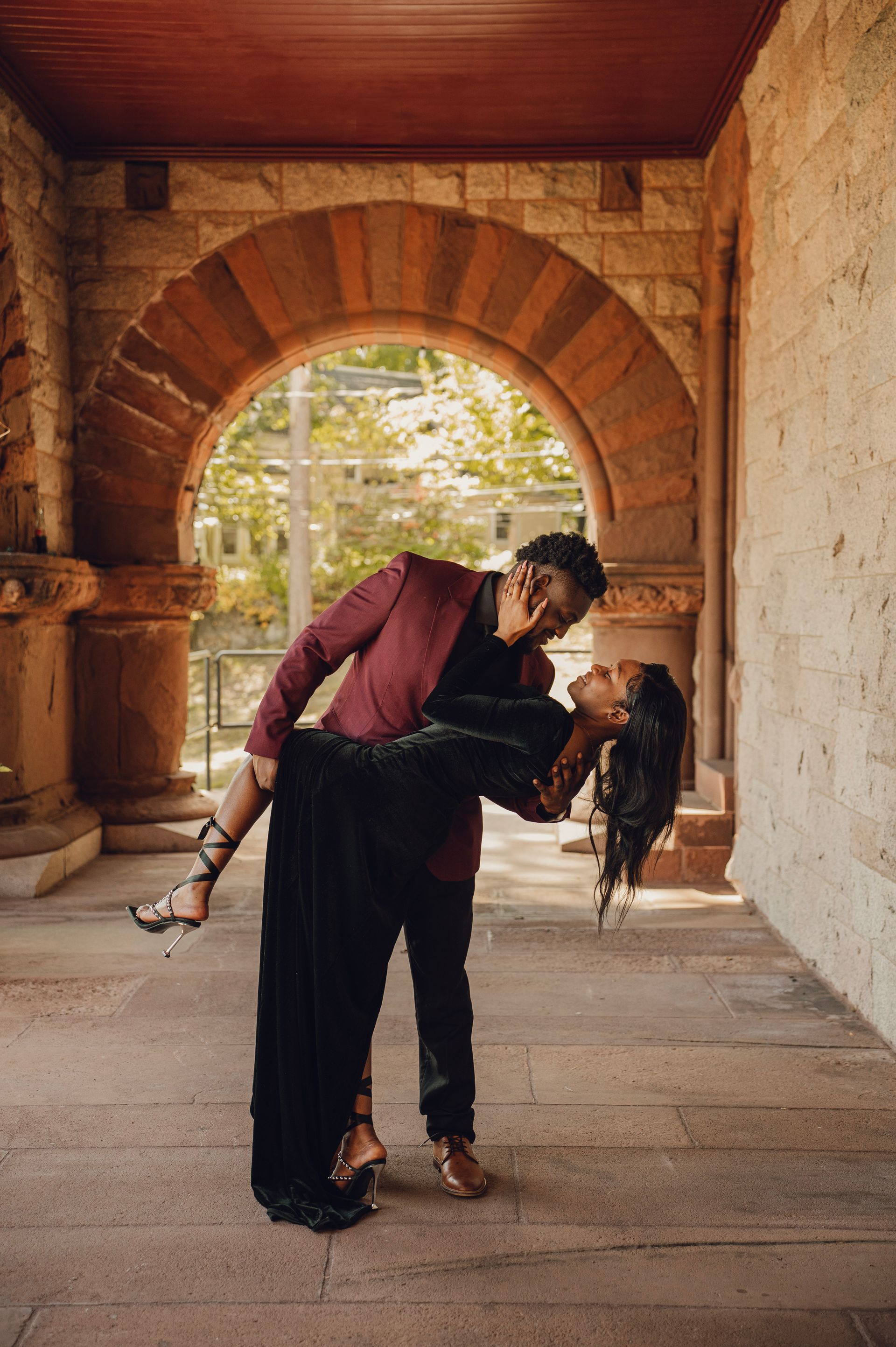 A person in a maroon blazer dips a person in a long black dress beneath a stone archway.