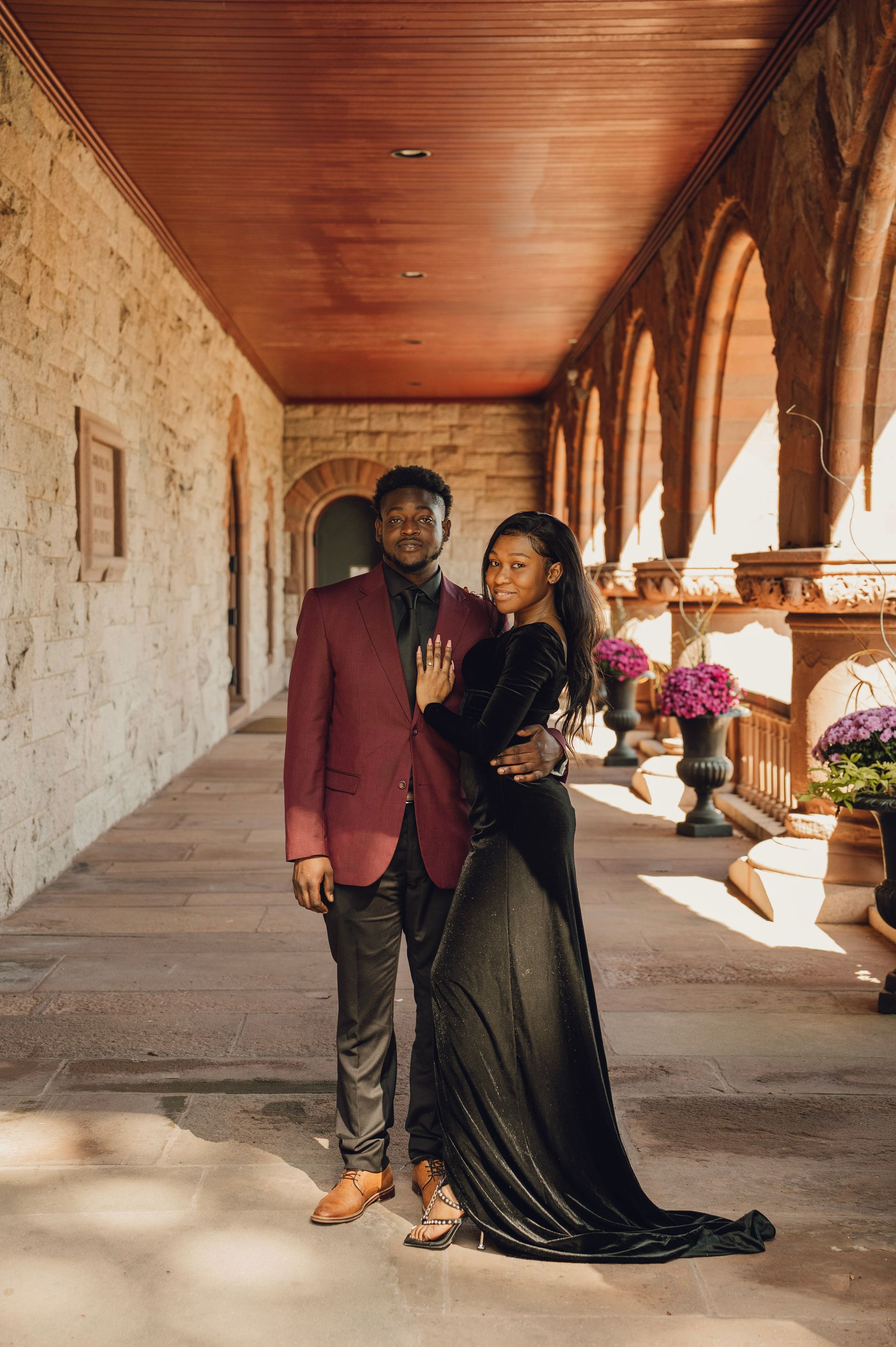 A couple stands together in a sunlit stone corridor with arched walkways, he in a maroon blazer, she in a long black dress.