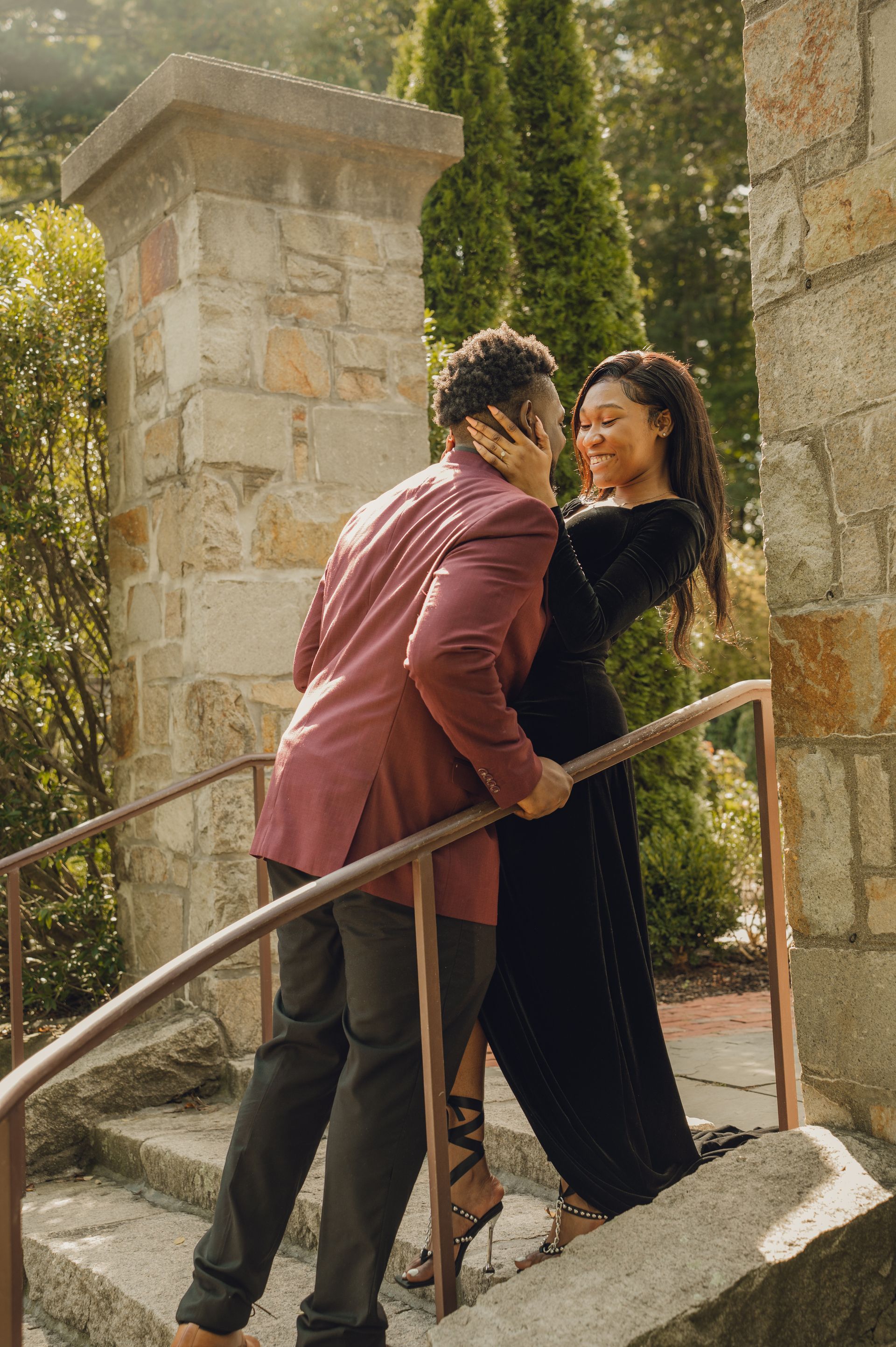 A couple in formal attire embraces on stone steps by a stone pillar, gazing lovingly at each other.