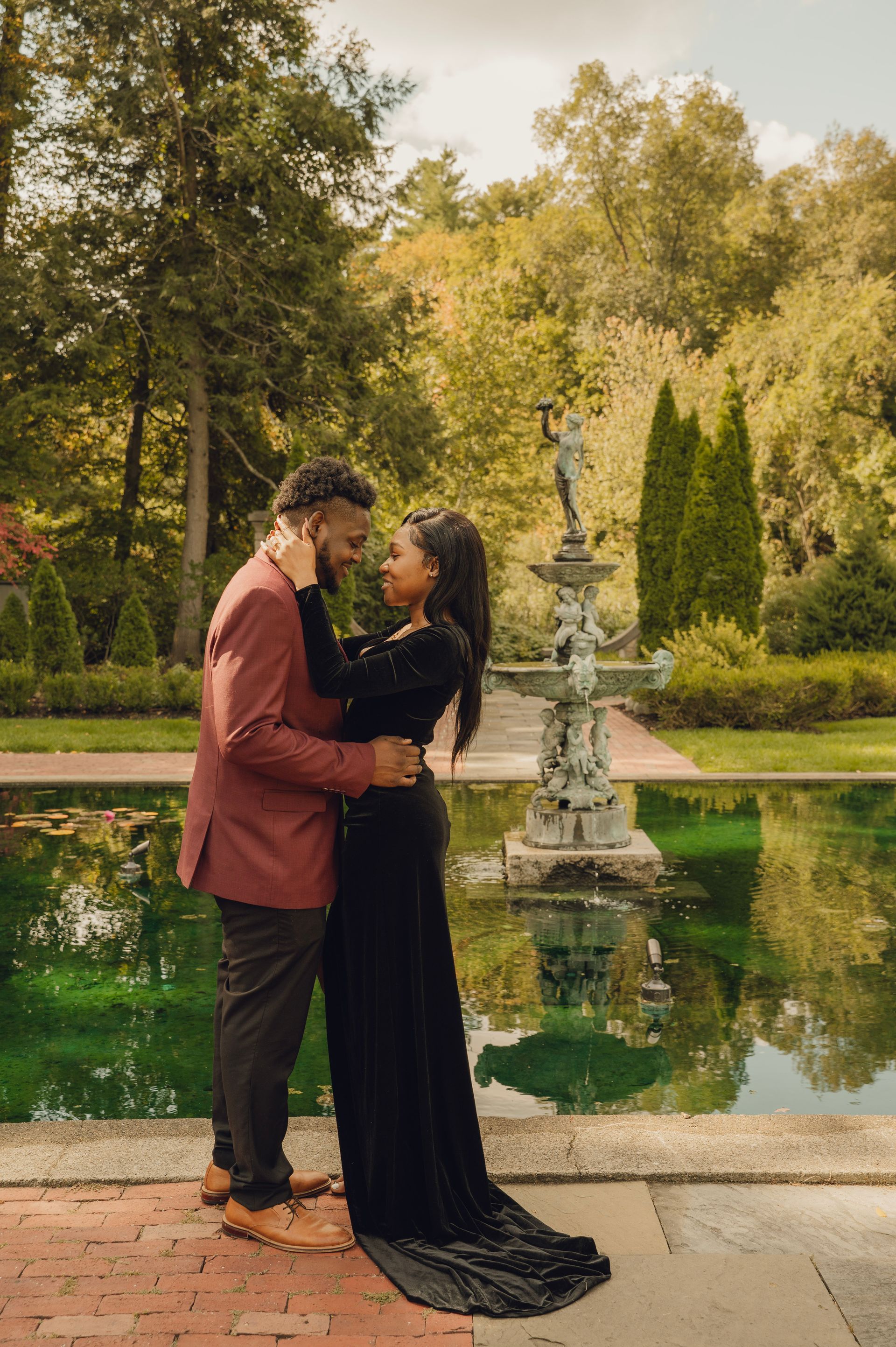 A couple standing face-to-face in a loving embrace beside a fountain in a lush garden.