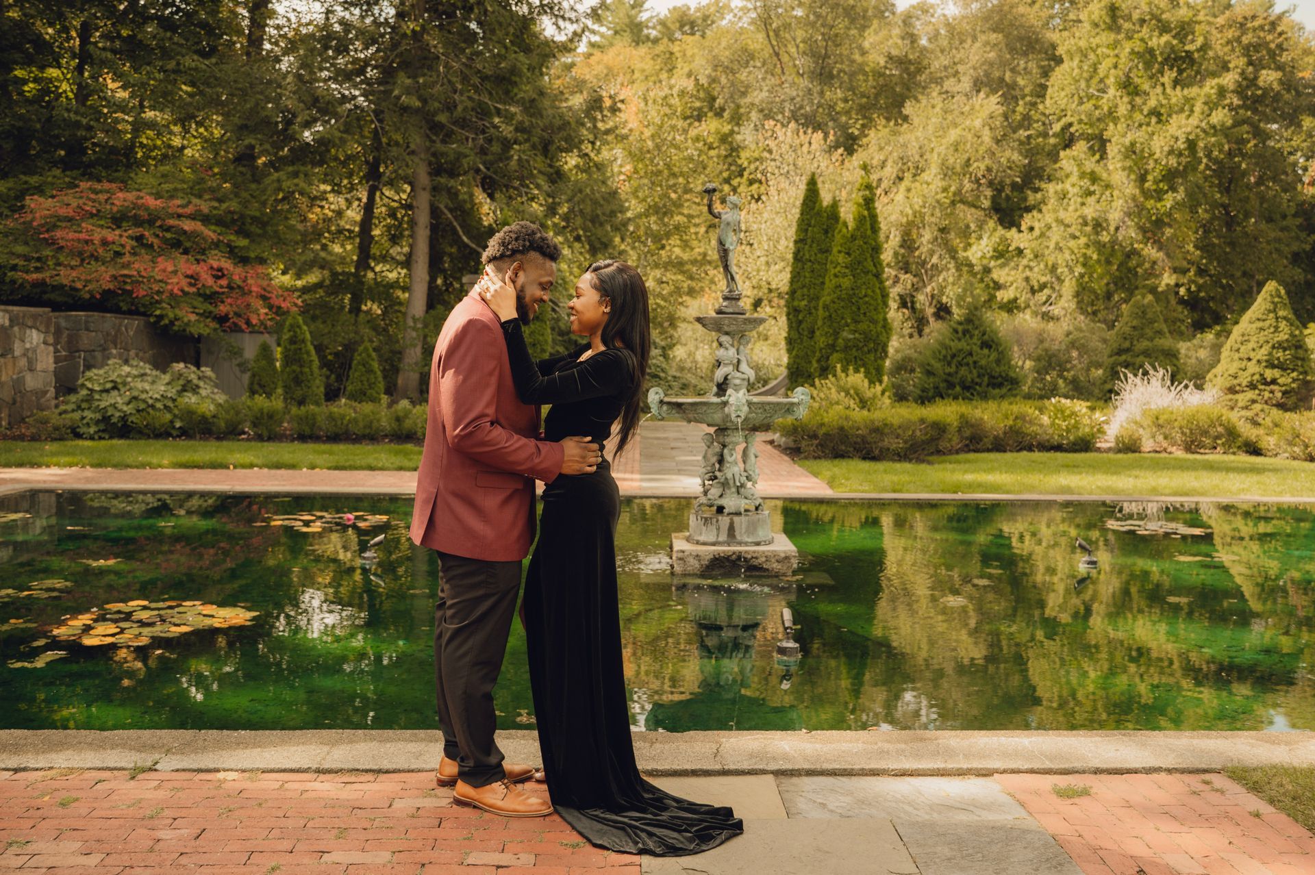 A couple in elegant attire embraces near a fountain in a lush, green garden pond setting.