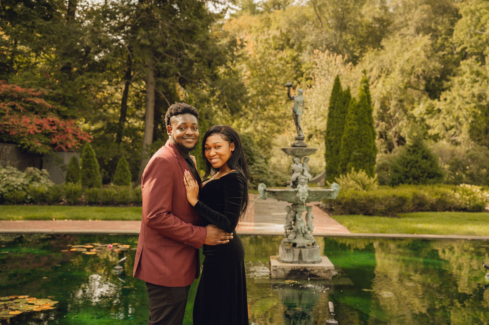 A couple in formal attire stands by a decorative fountain in a lush, green garden, posing together for a portrait.