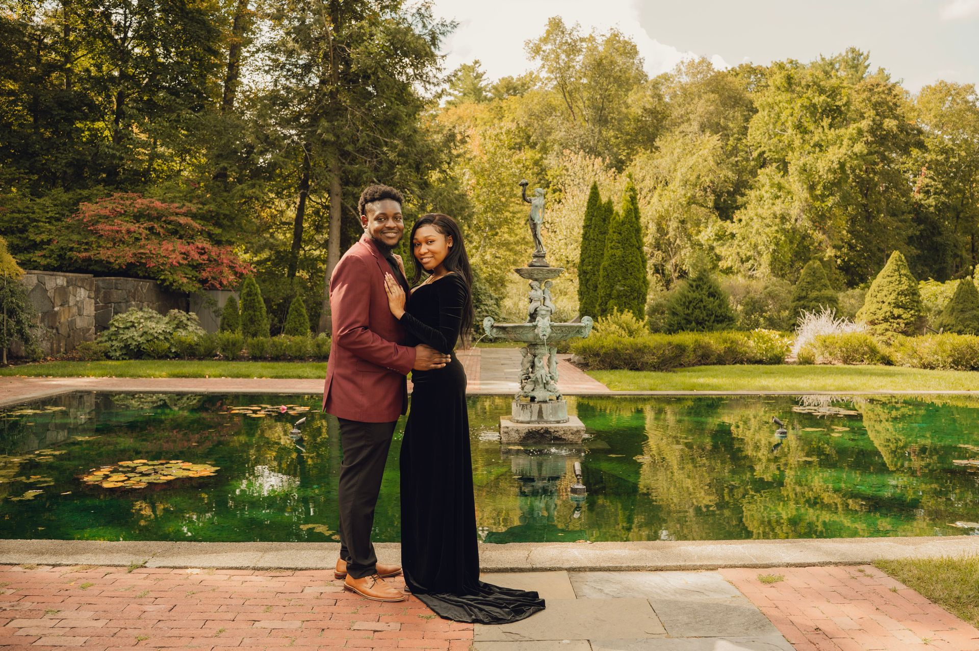 A couple stands arm-in-arm posing before a decorative garden fountain and pond on a sunny day.