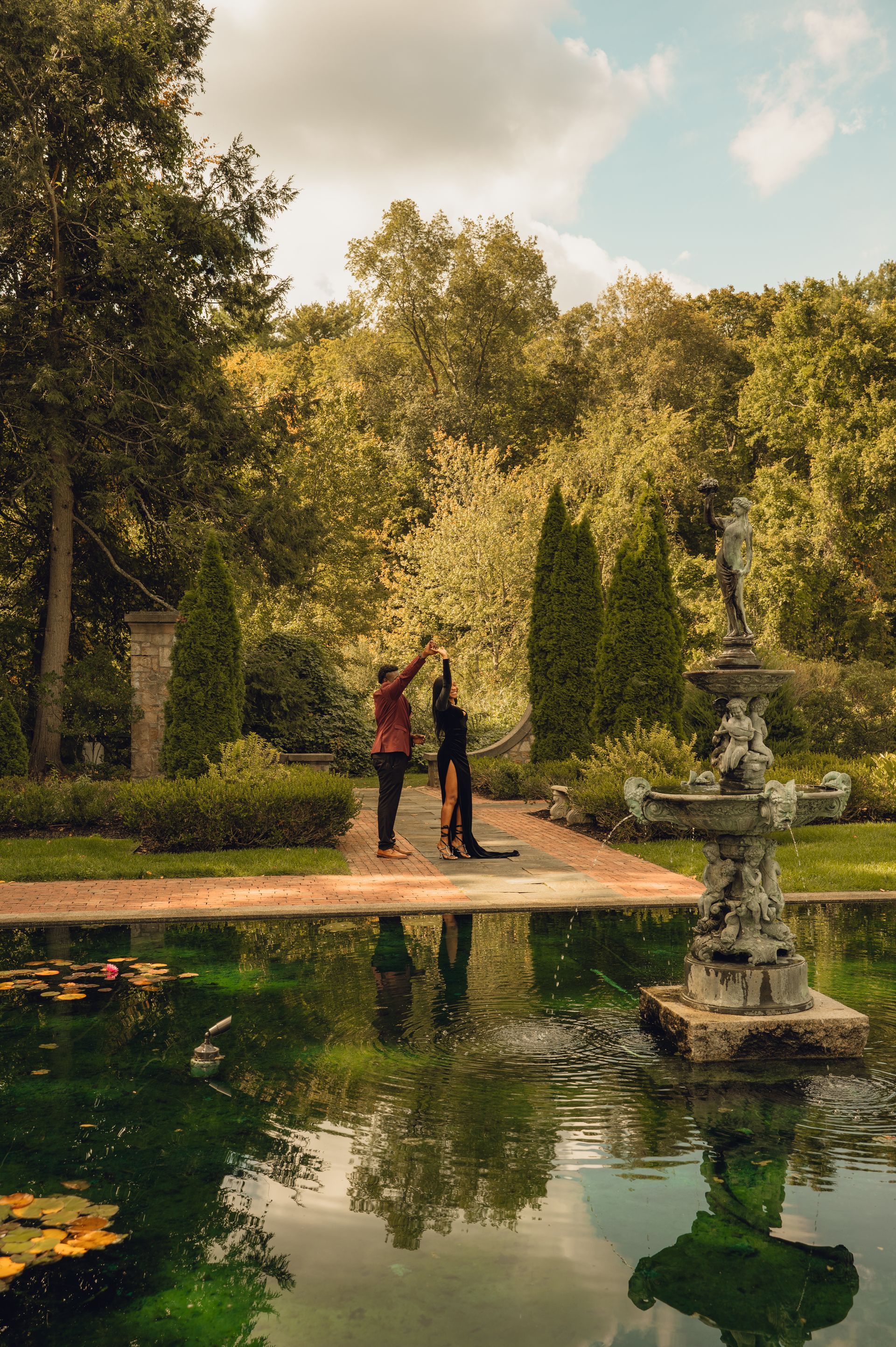 Two people stand on a path in a lush garden by a pond, one reaching up toward tree branches near a stone fountain.