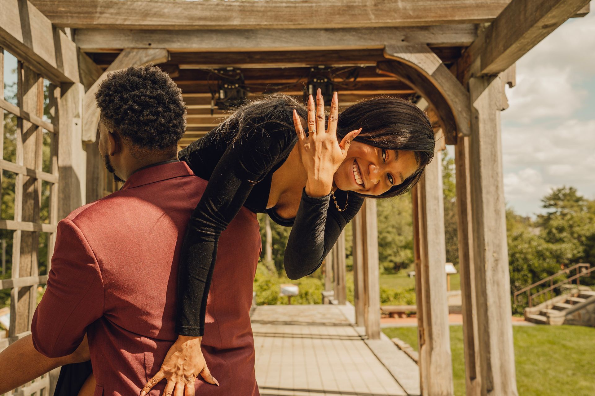 A person in a black dress is being held by a partner in a red shirt, smiling and showing off a ring on their finger.