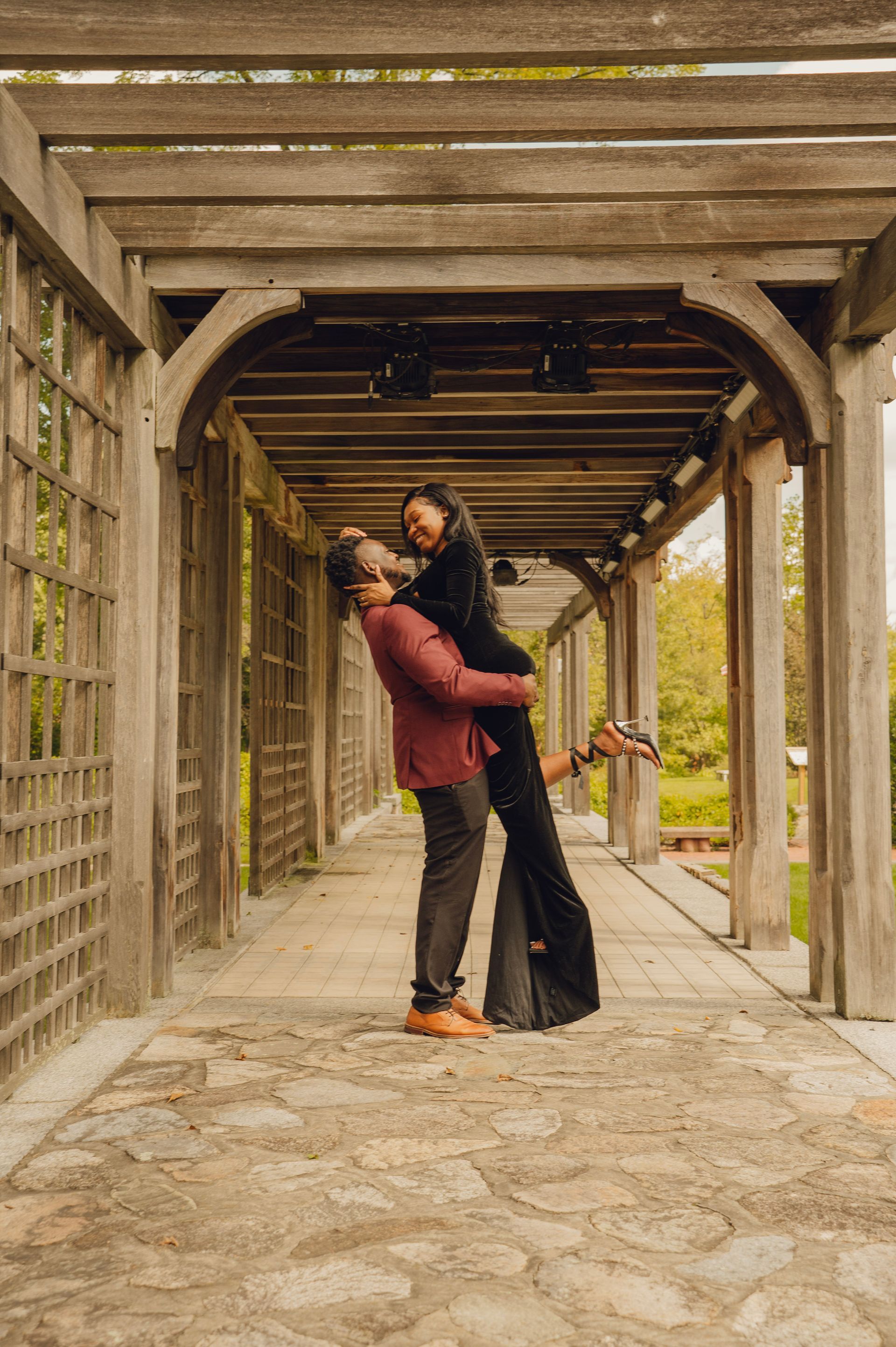 A person in a maroon jacket lifts another in a black gown inside a wooden pergola.