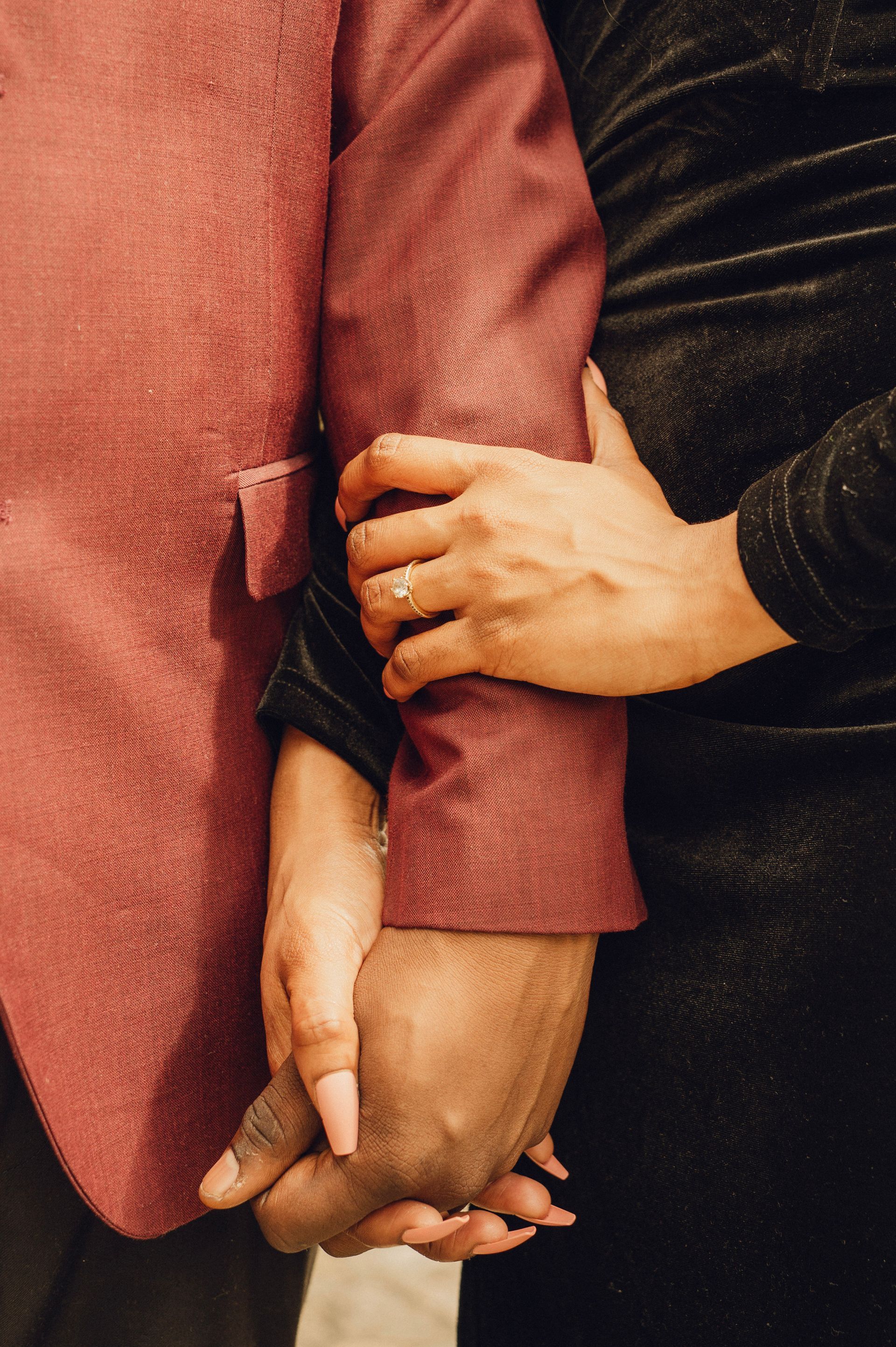 Two hands holding each other; one wears a ring, with a burgundy sleeve on the left and a black sleeve on the right.