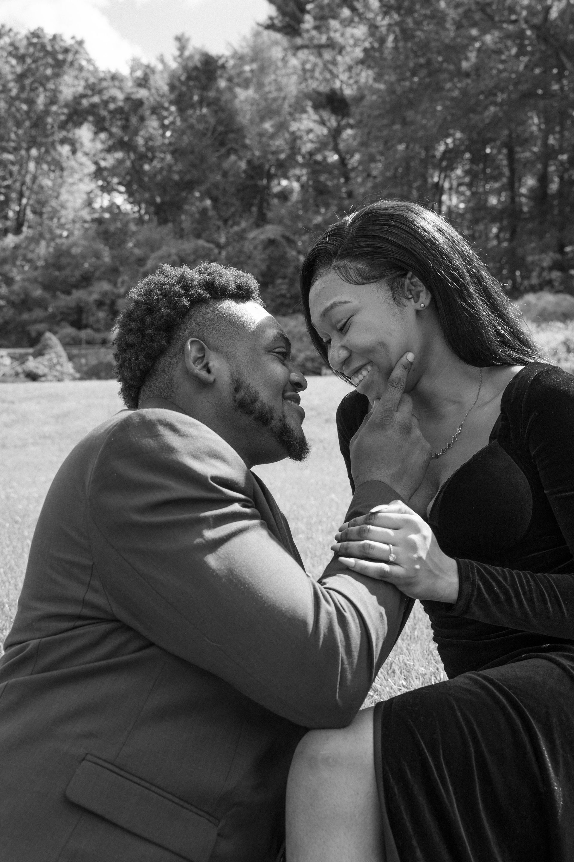 A person in a suit kneels before someone in a dress, gently touching their face in a field outdoors.