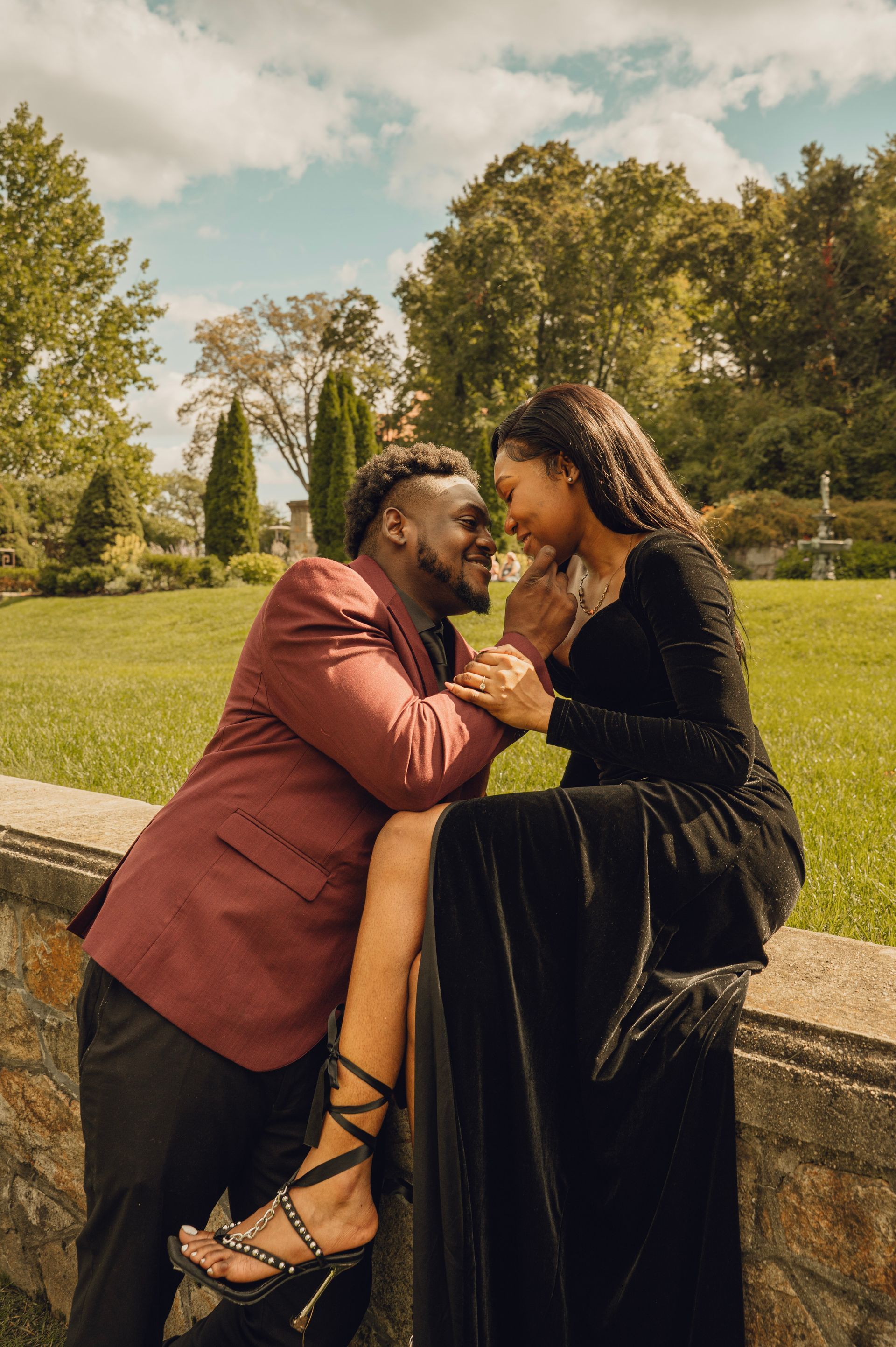 A couple in elegant attire poses intimately by a stone wall in a sunlit grassy field.