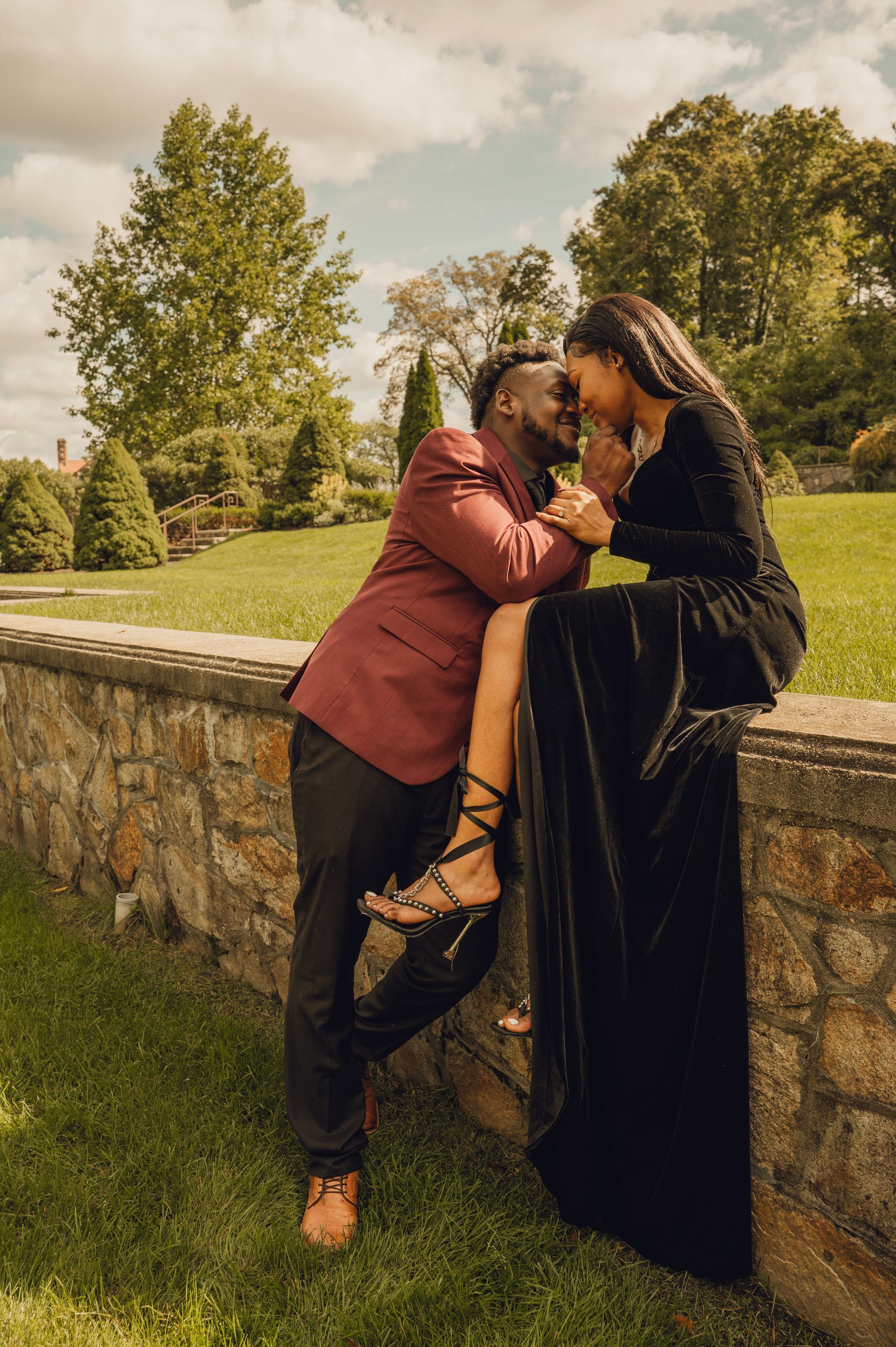 A couple in formal attire embraces while sitting and standing on a stone wall in a sunlit grassy landscape.