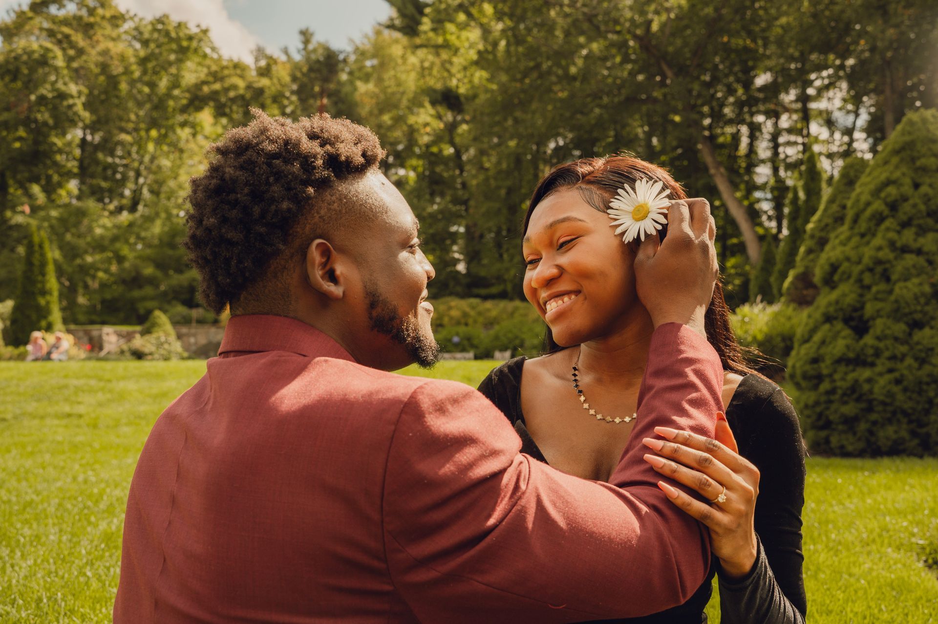 A person in a maroon jacket tucks a white daisy behind the ear of a person in a black dress in a sunlit garden.