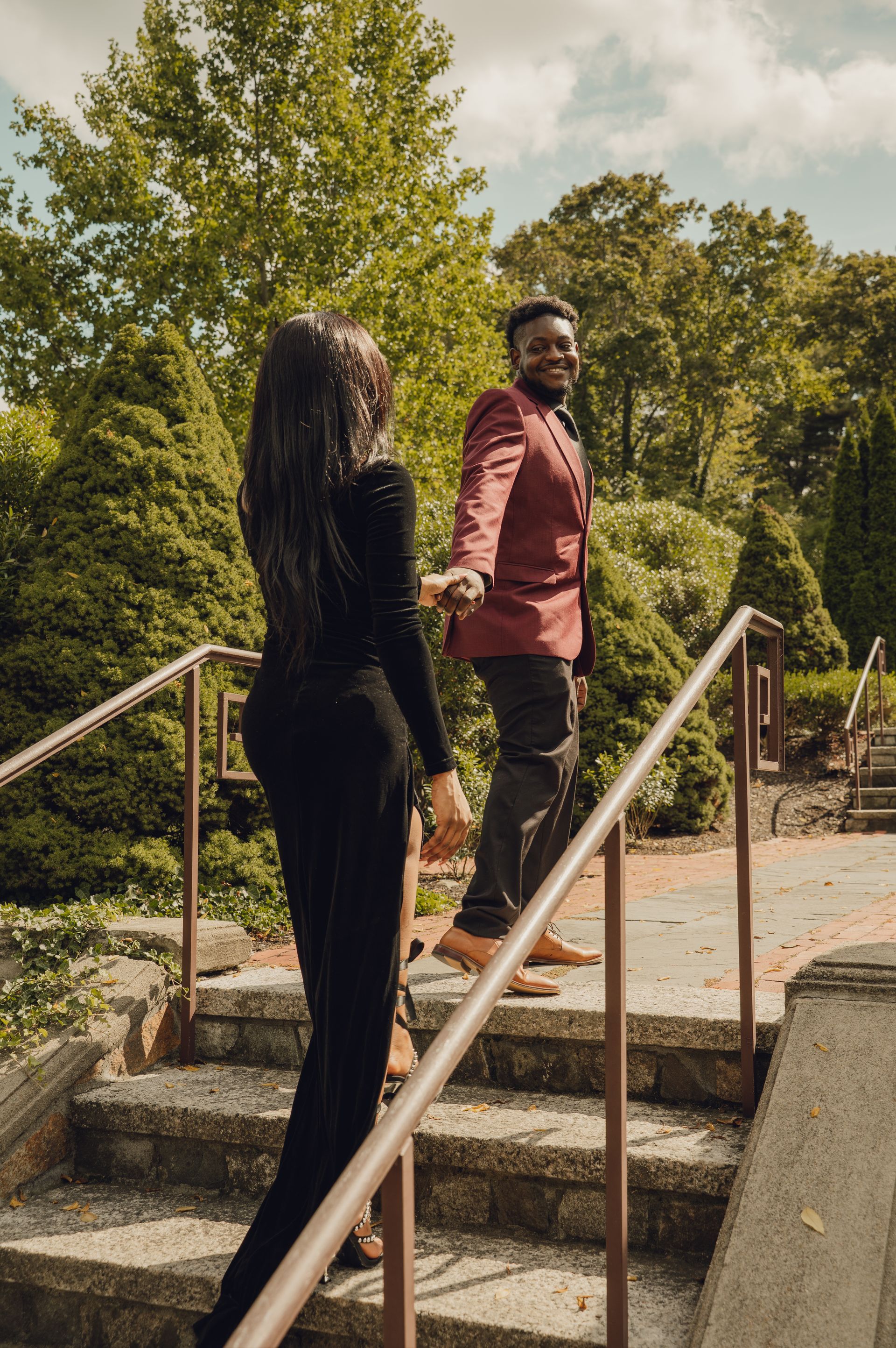 A person in a red suit leads another in a black outfit up outdoor stone stairs in a park.