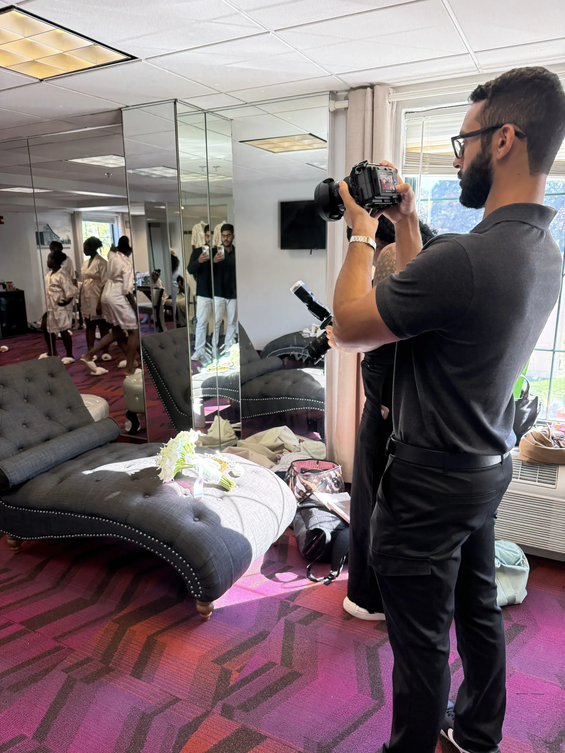 A photographer captures a group in robes through a large mirror in a room with a gray chaise lounge and purple carpet.