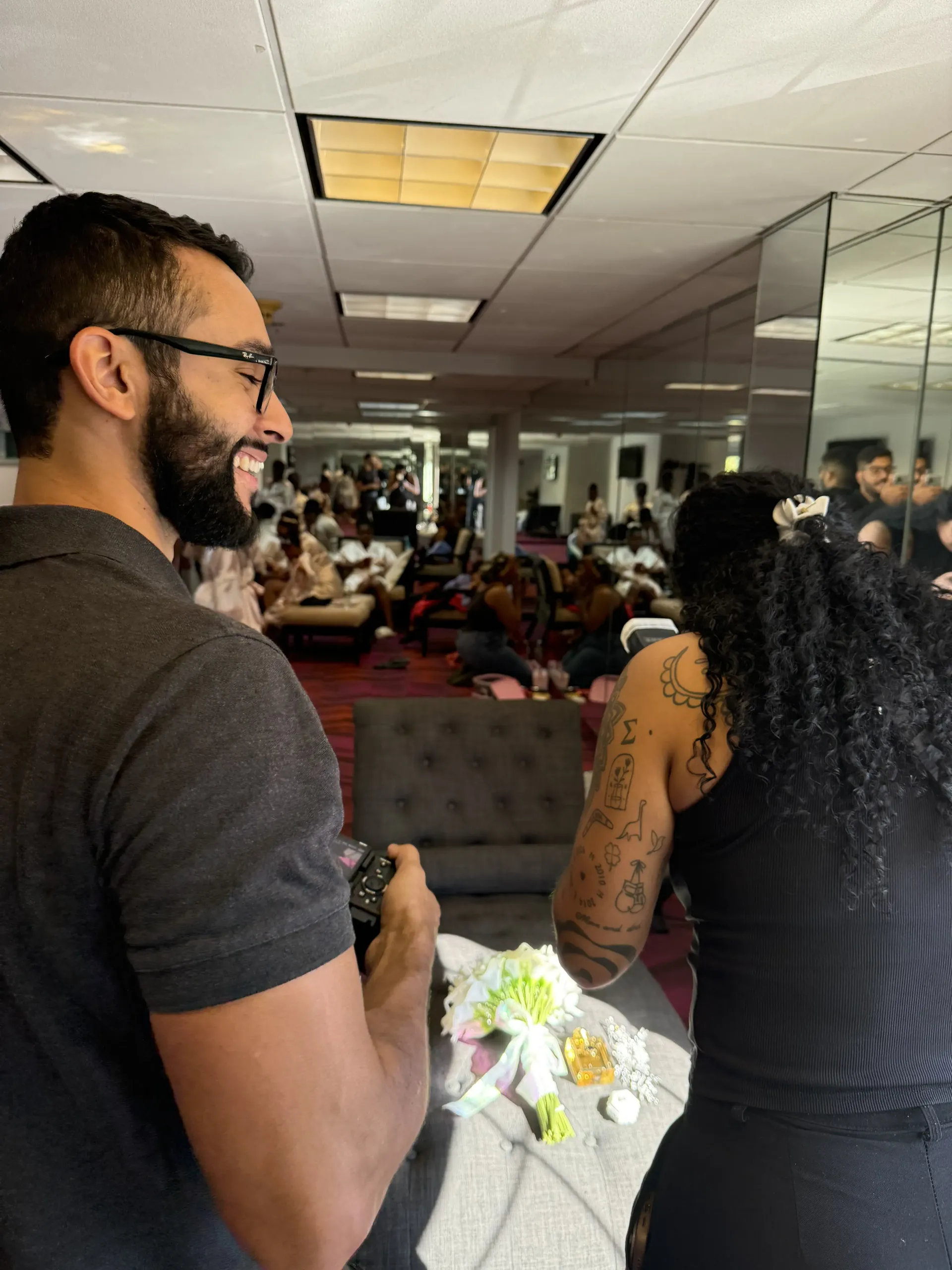 A man with glasses and a beard smiling while holding a camera, standing near a person with a tattoo on their arm.