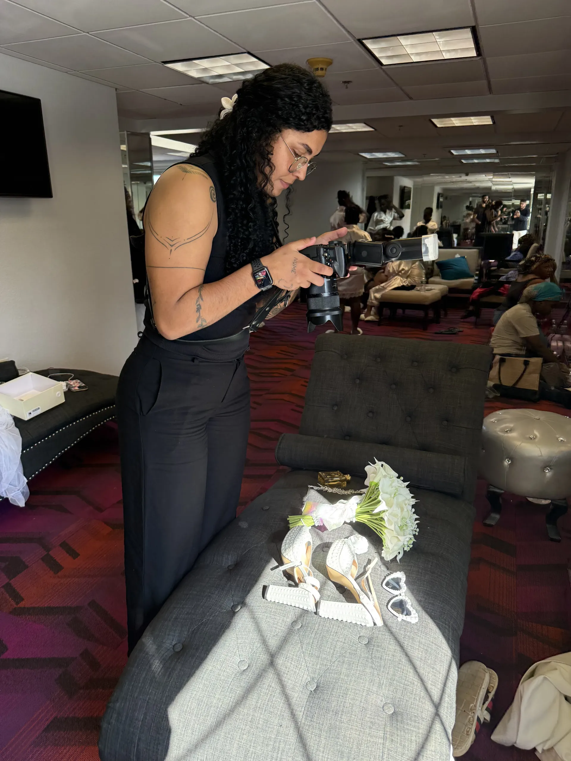 A person with long curly hair uses a camera to photograph bridal accessories arranged on a tufted chaise lounge.