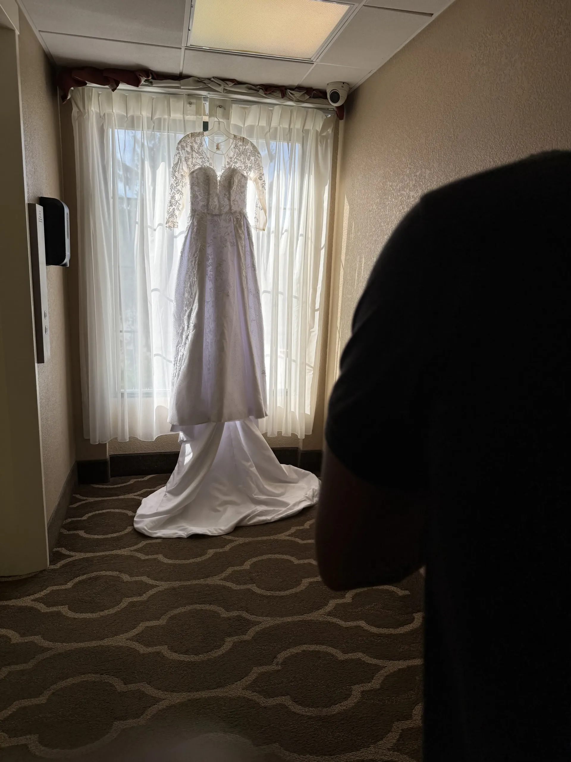 A white wedding dress hangs against a draped window in a room with patterned carpet, with a person partially in view.