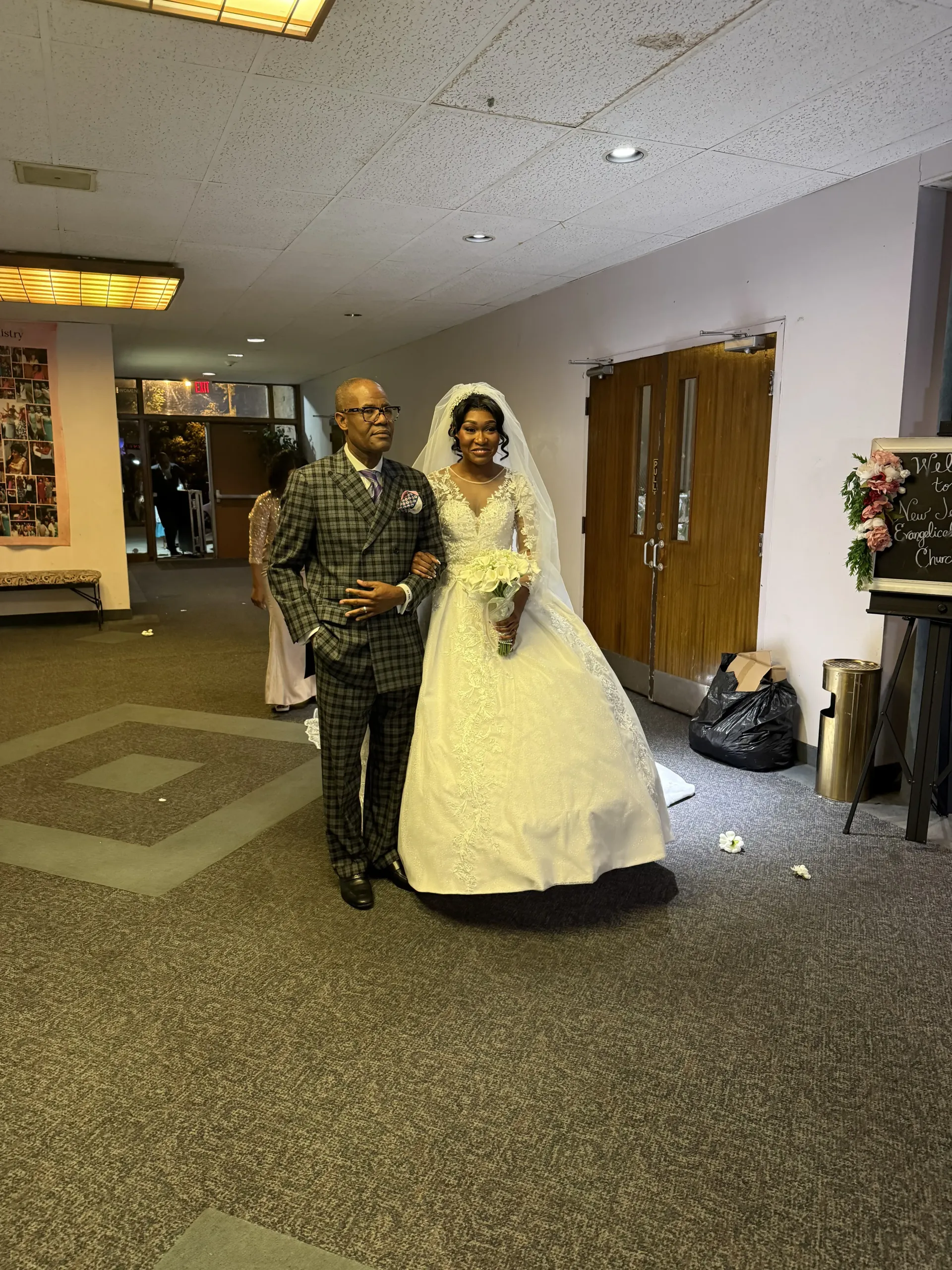 A person in a patterned suit walks a person in a white wedding gown and veil holding a bouquet through a hallway.