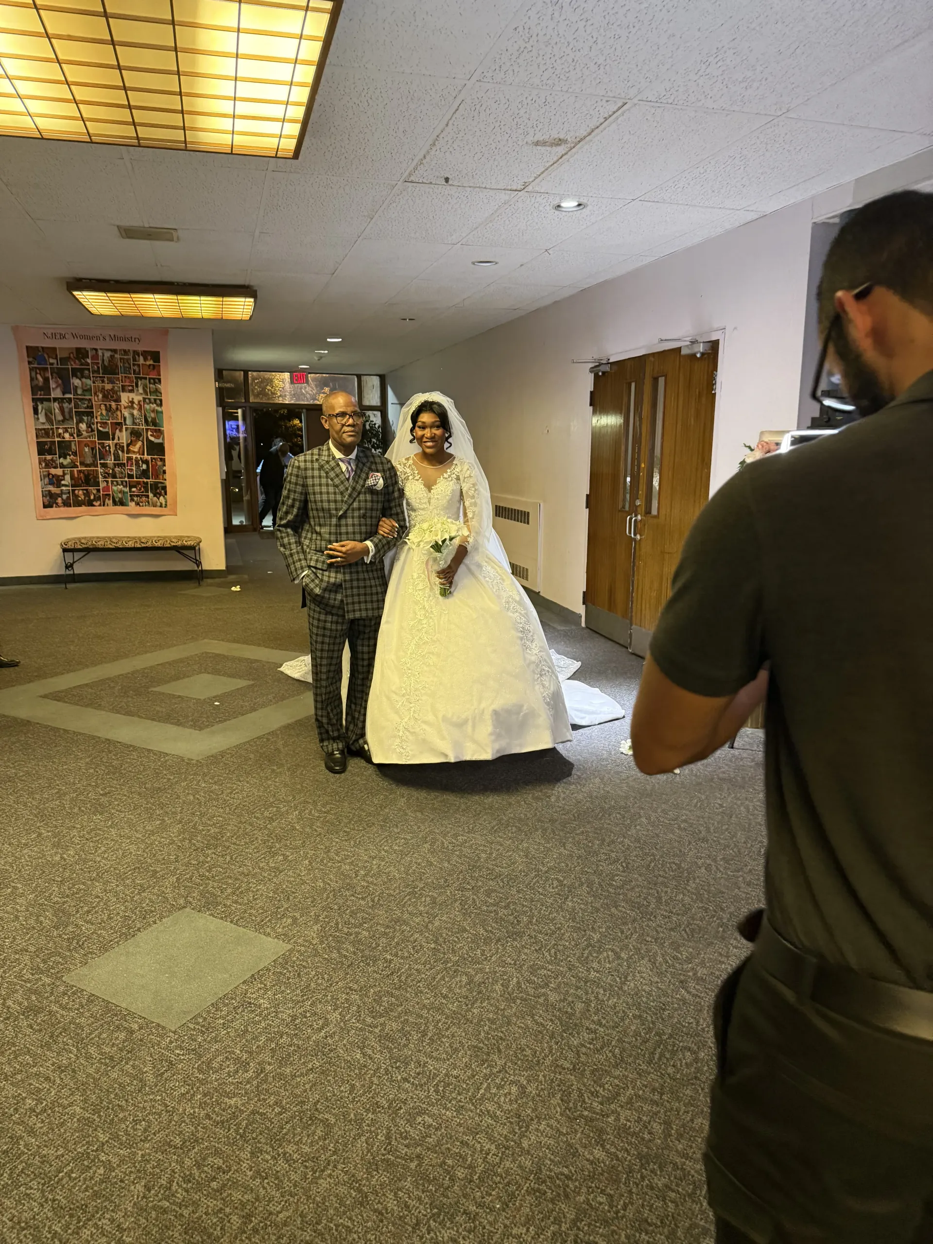 A person in a wedding dress and a person in a suit walk down a hallway toward a camera held by a photographer.