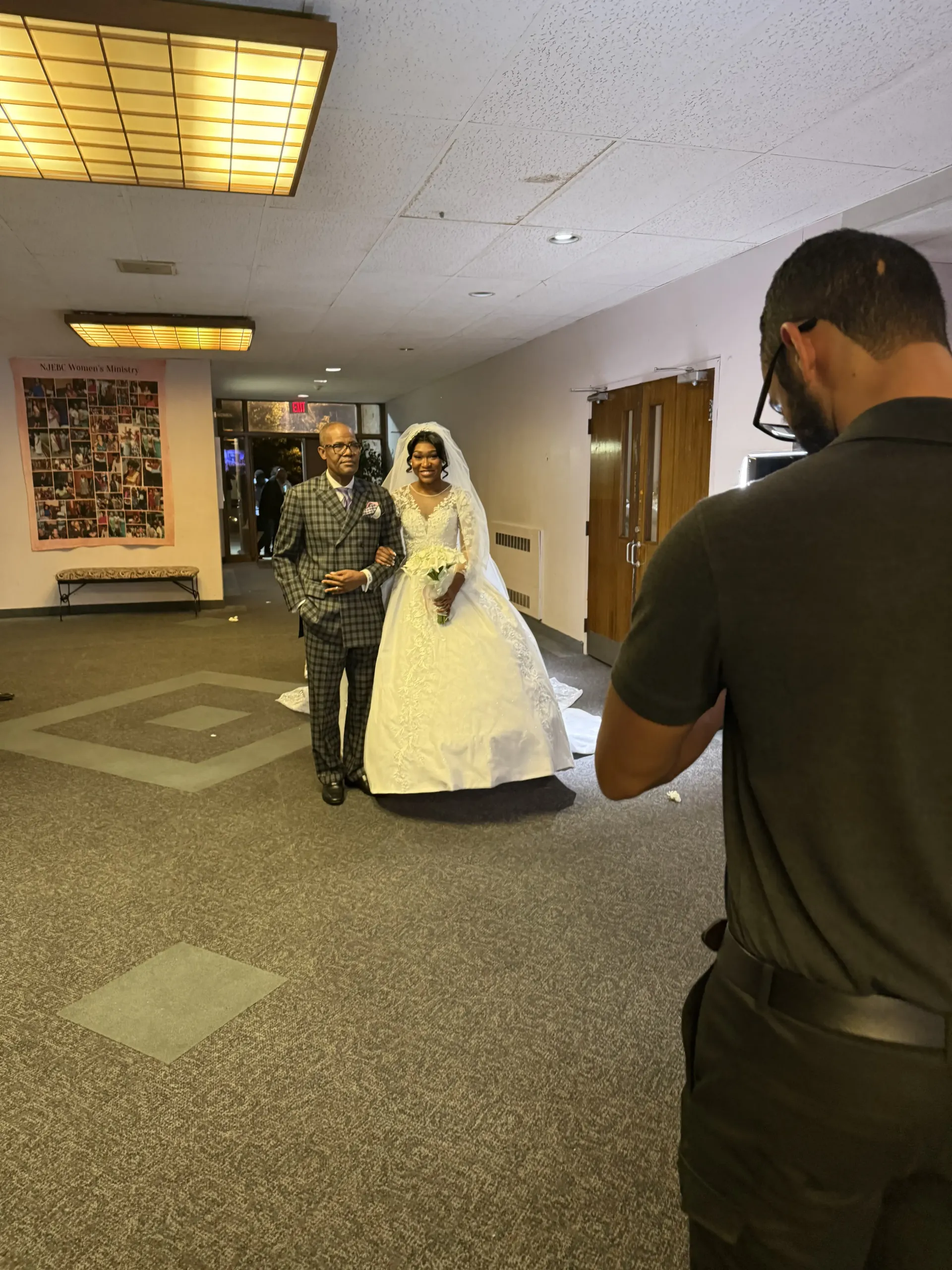 A photographer films a smiling bride in a white gown and a man in a patterned suit walking through a hallway.