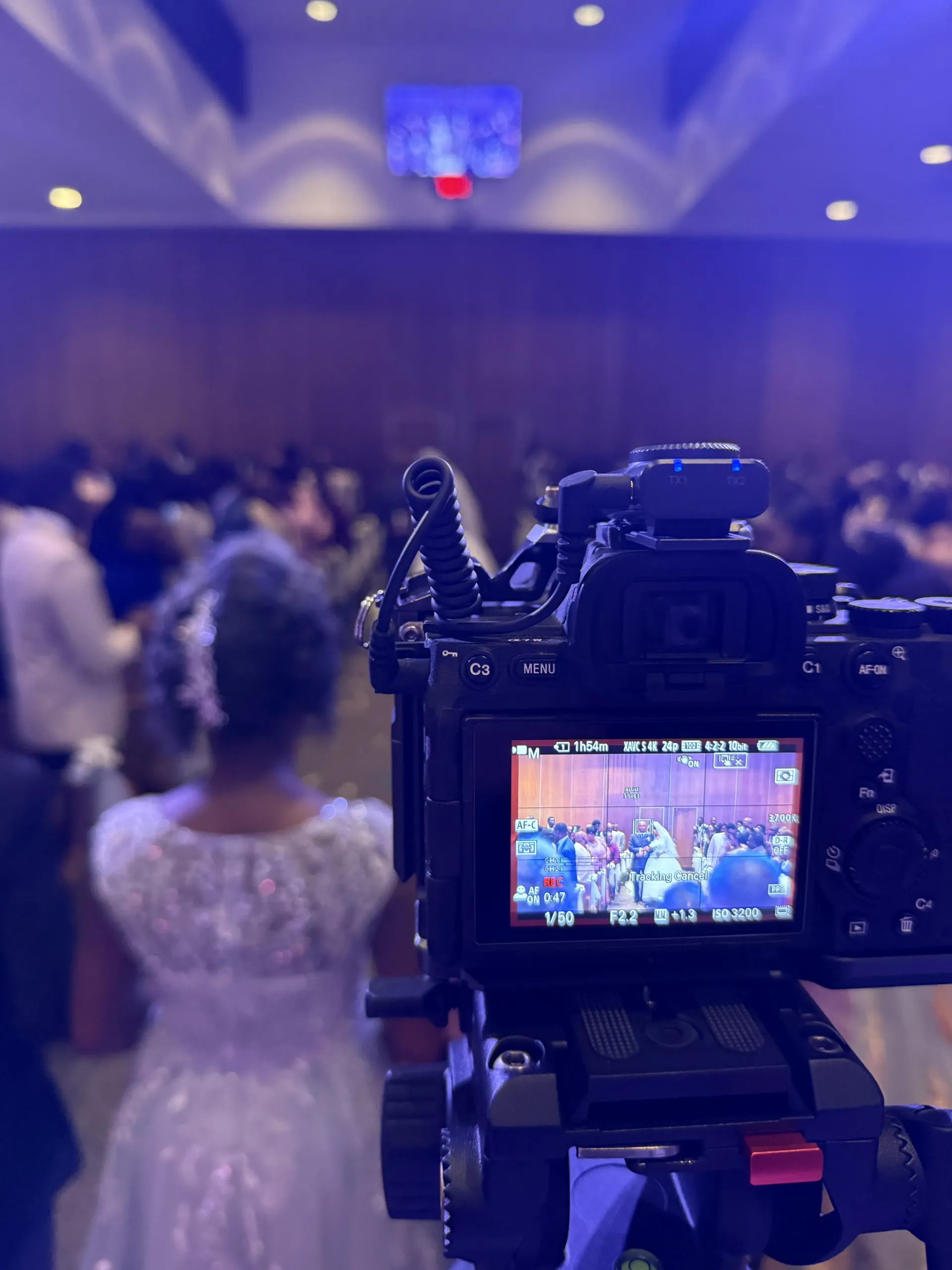 A camera on a tripod faces a wedding ceremony, showing the event on its screen with a person in a white dress in view.