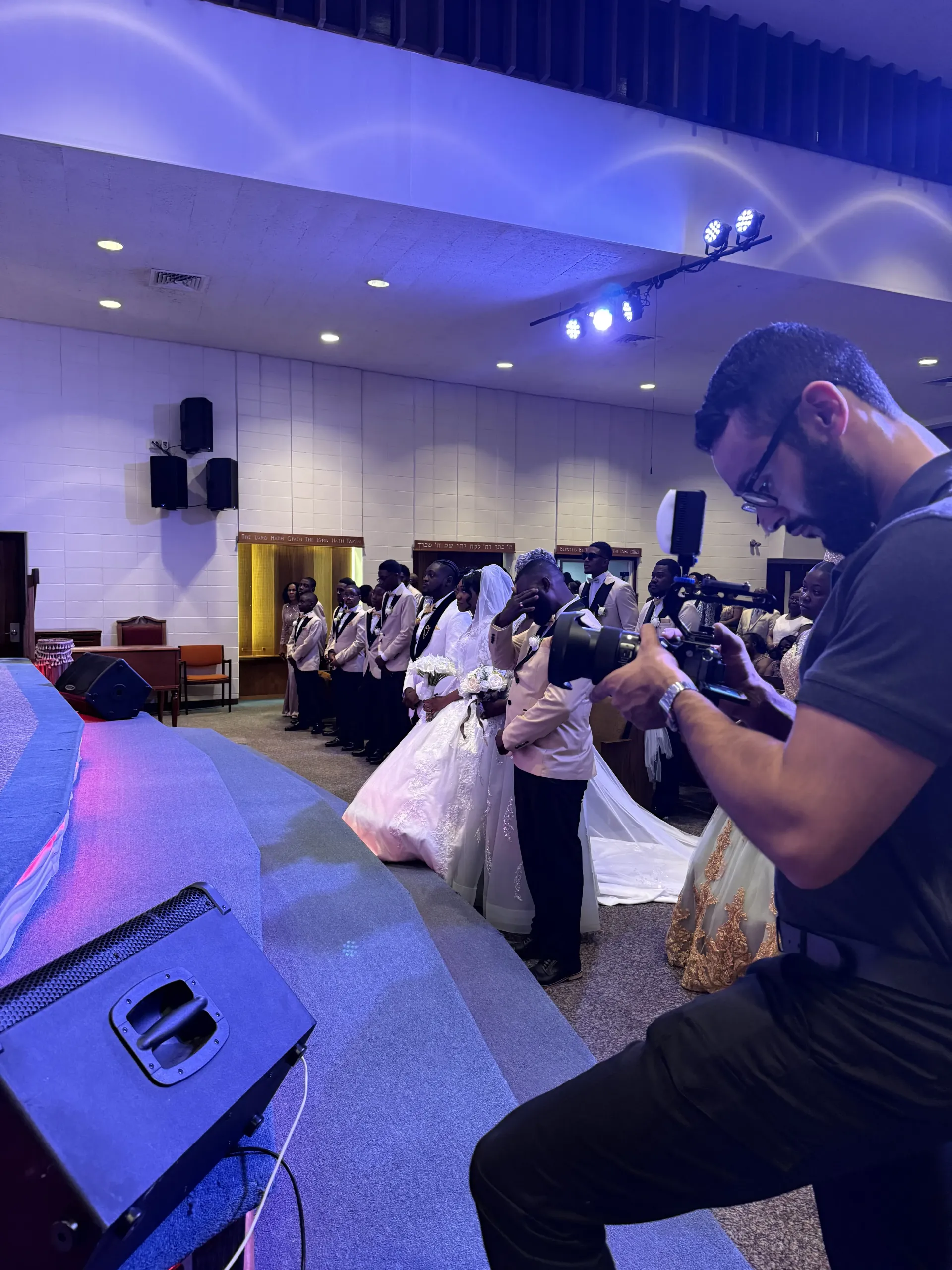 A videographer films a wedding procession inside a dimly lit venue with blue stage lighting and a purple carpeted floor.