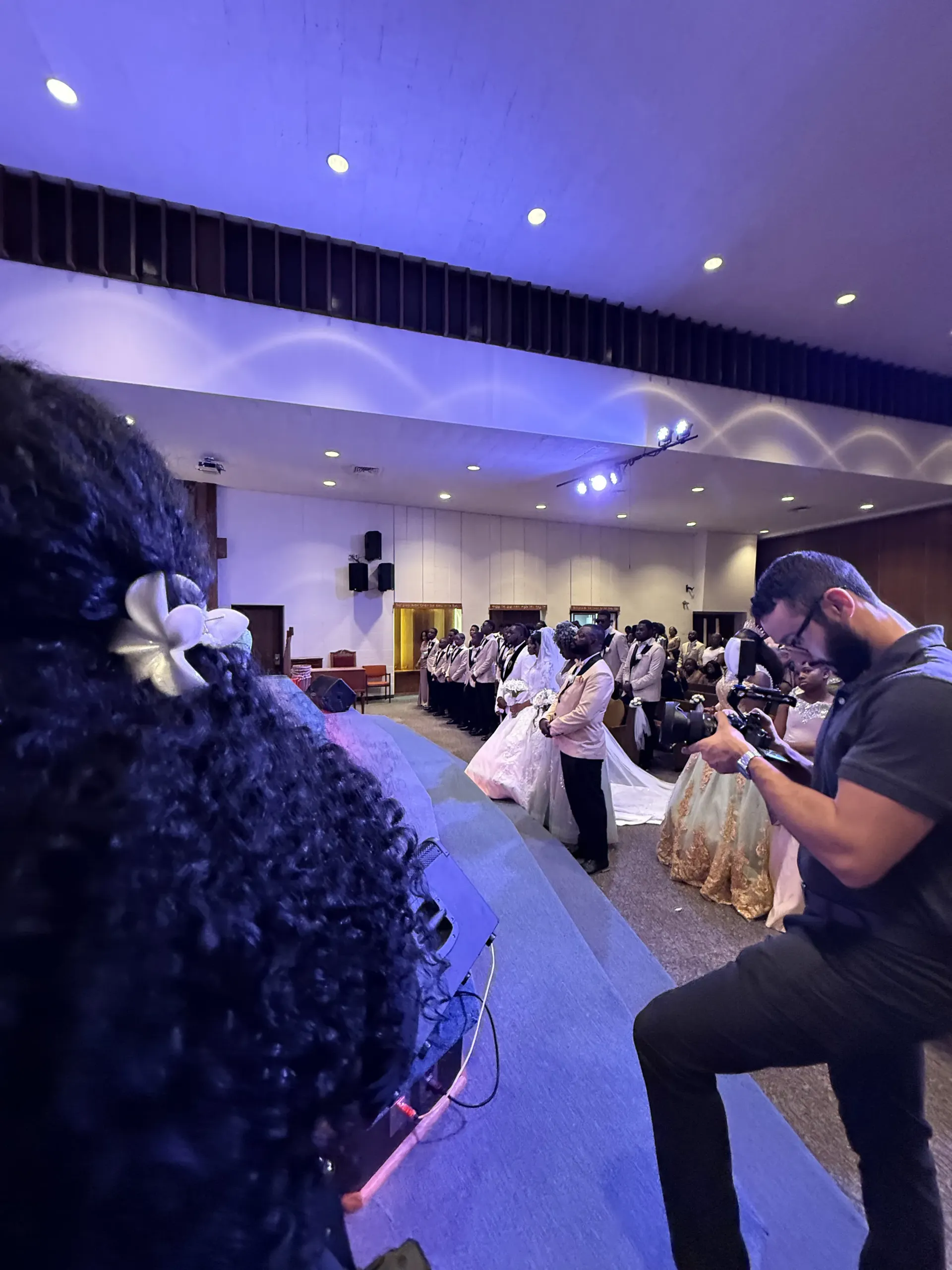 A photographer captures a wedding ceremony from the side as guests stand in a large, dimly lit hall.