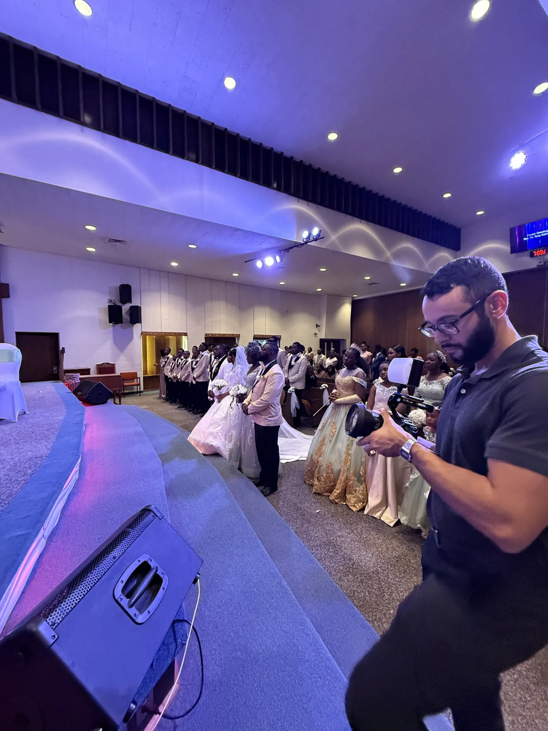 A videographer films a wedding party standing on a stage in a large, brightly lit indoor auditorium.
